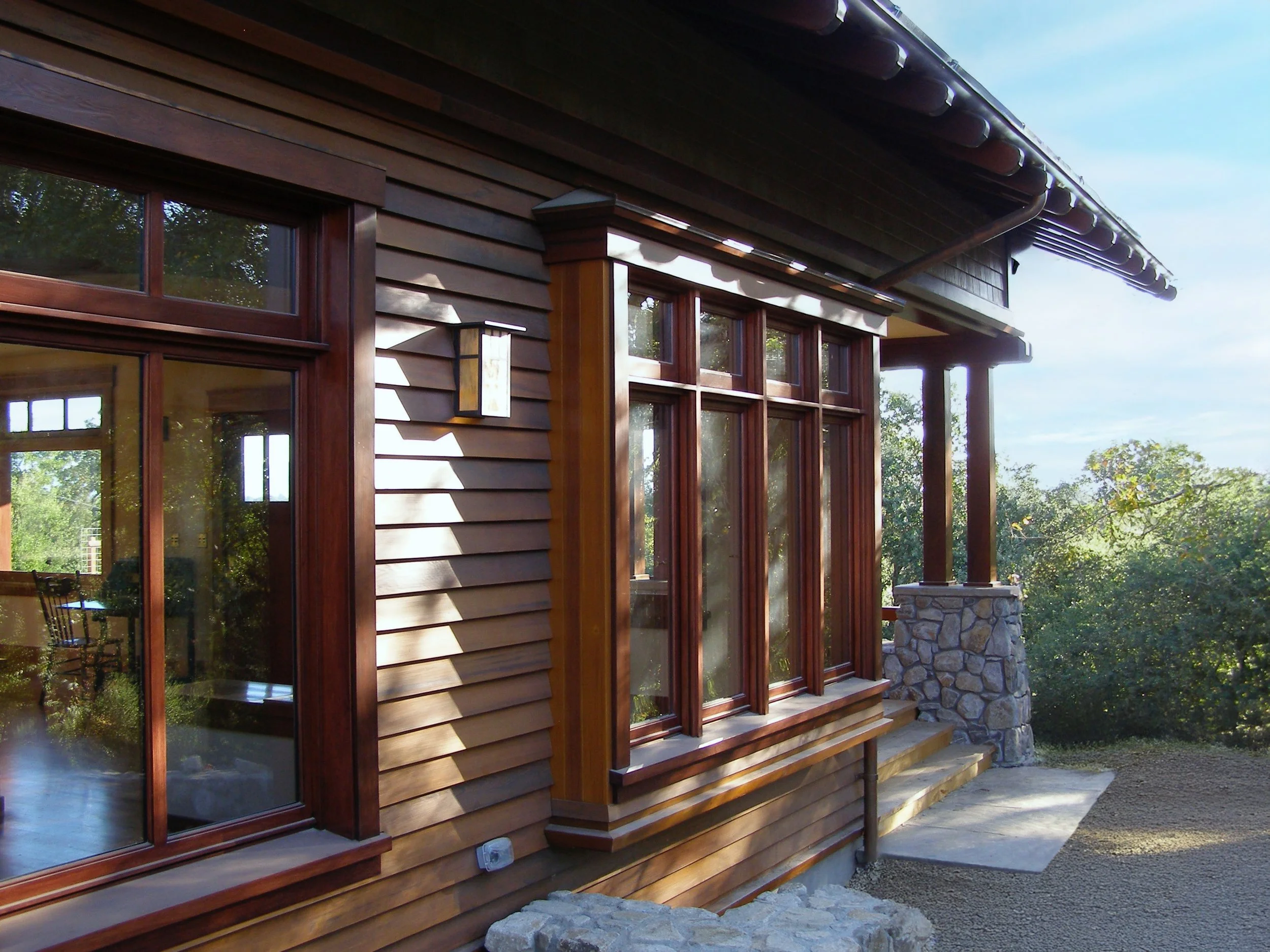 Exterior of a house with wooden siding, large windows, a stone chimney, and a metal roof.