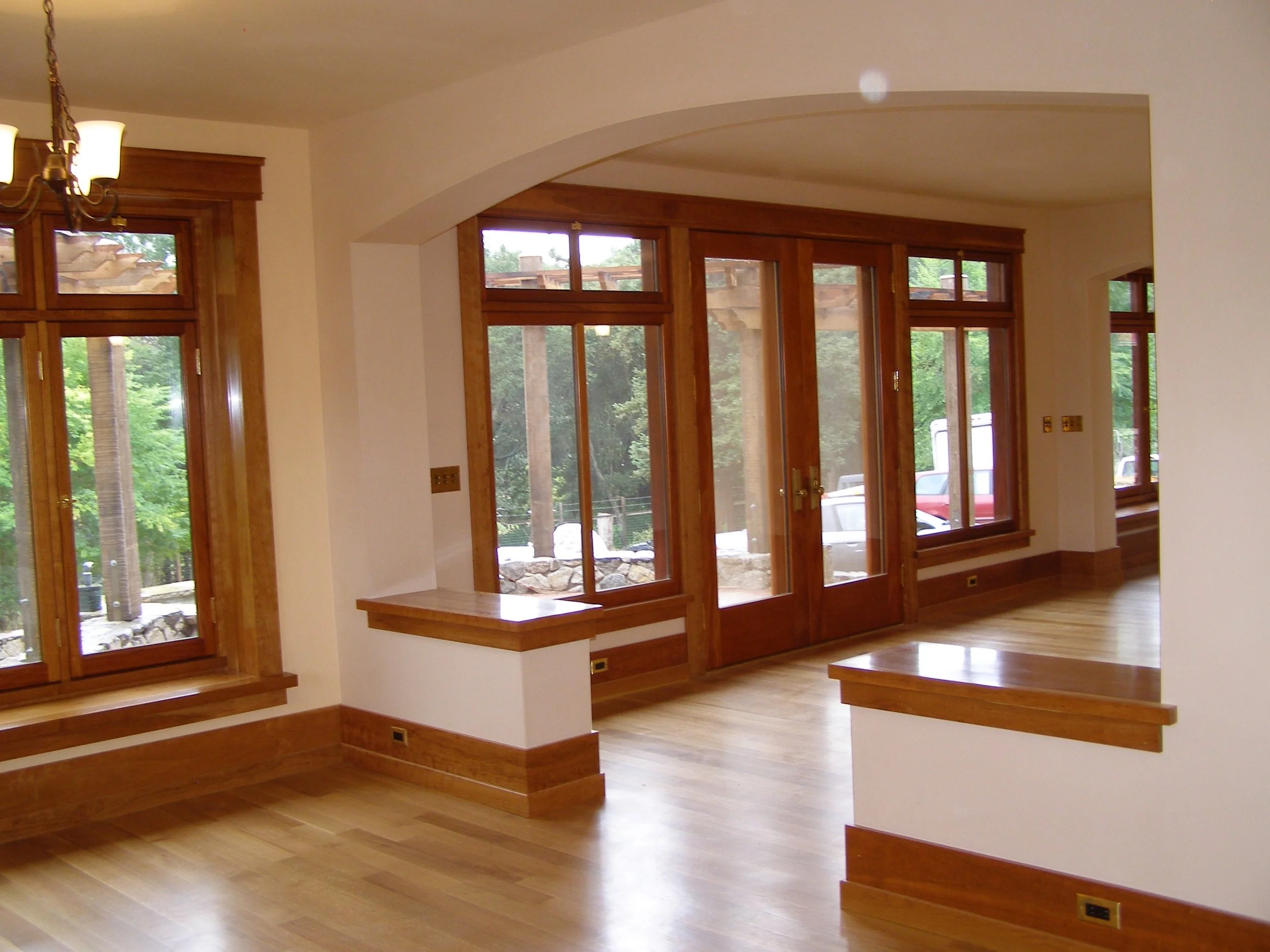 A spacious living room with large wooden framed windows allowing natural light, hardwood floors, and a wooden ledge or counter along the windows. A chandelier with multiple lamps hangs from the ceiling.