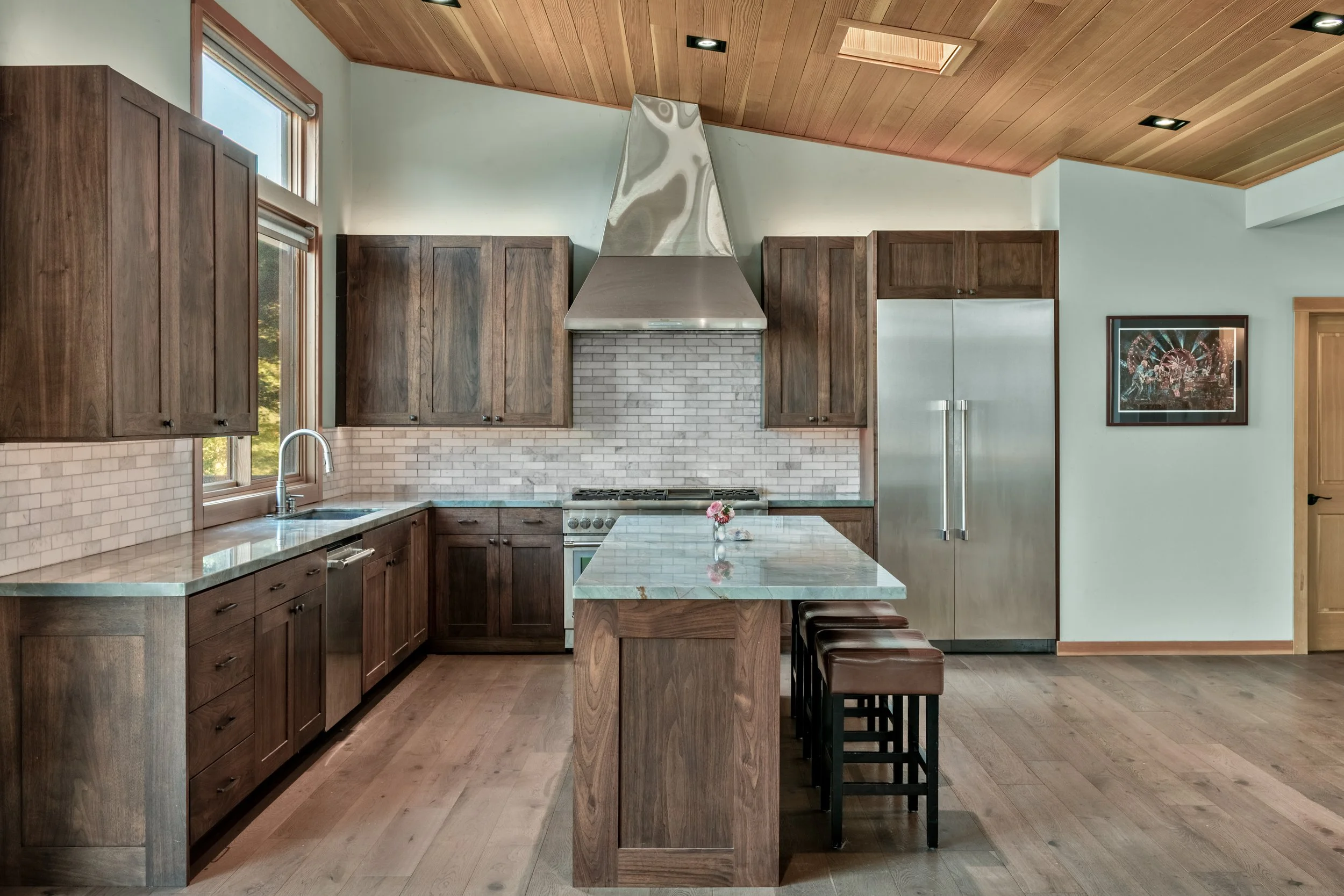 Modern kitchen with wooden cabinets, marble countertops, a stainless steel range hood, and a small island with bar stools.