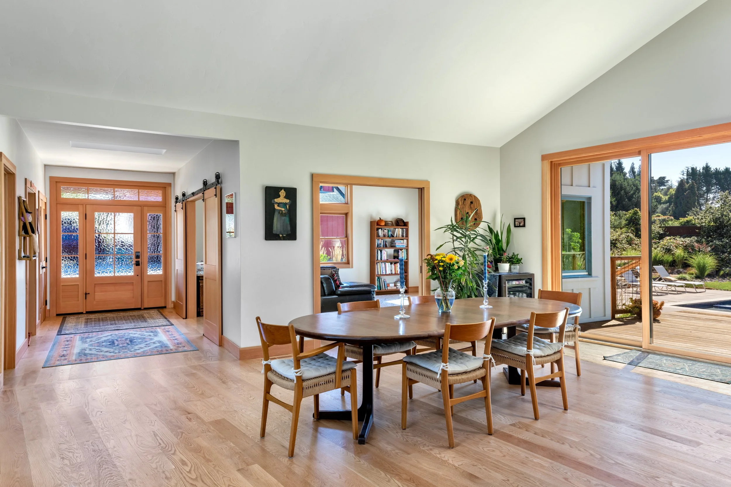 Living room with a wooden dining table and chairs, large windows and sliding glass door leading to a deck with outdoor furniture, plants, artwork on the walls, and hardwood flooring.