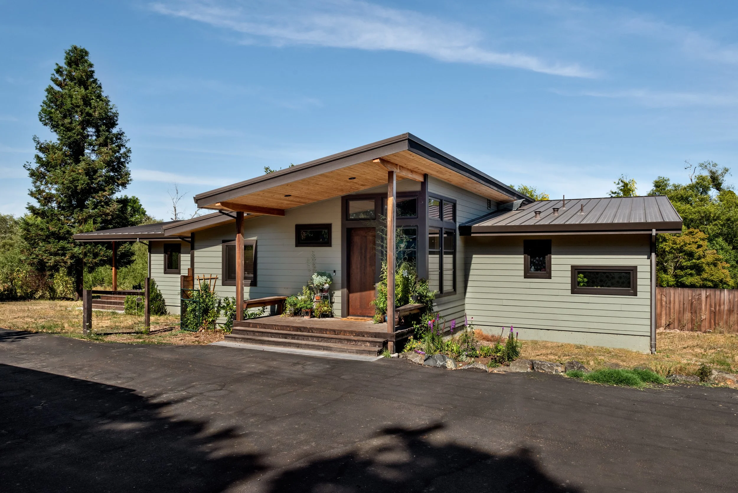 Modern house with a sloped roof and large windows, surrounded by trees and a small garden, under a clear blue sky.