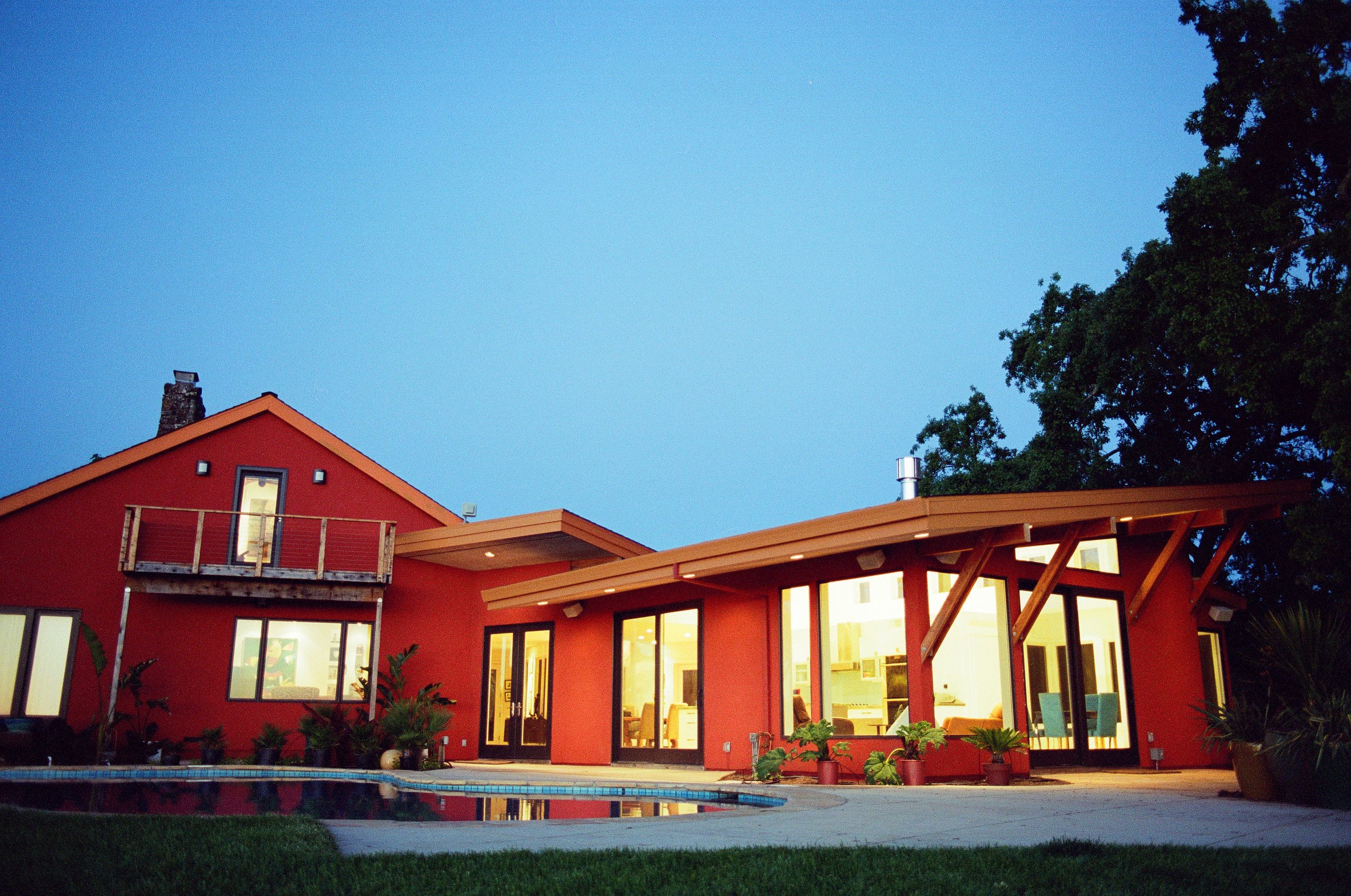 Red house with large windows, balcony, and outdoor patio with swimming pool at dusk, neighboring trees in the background.