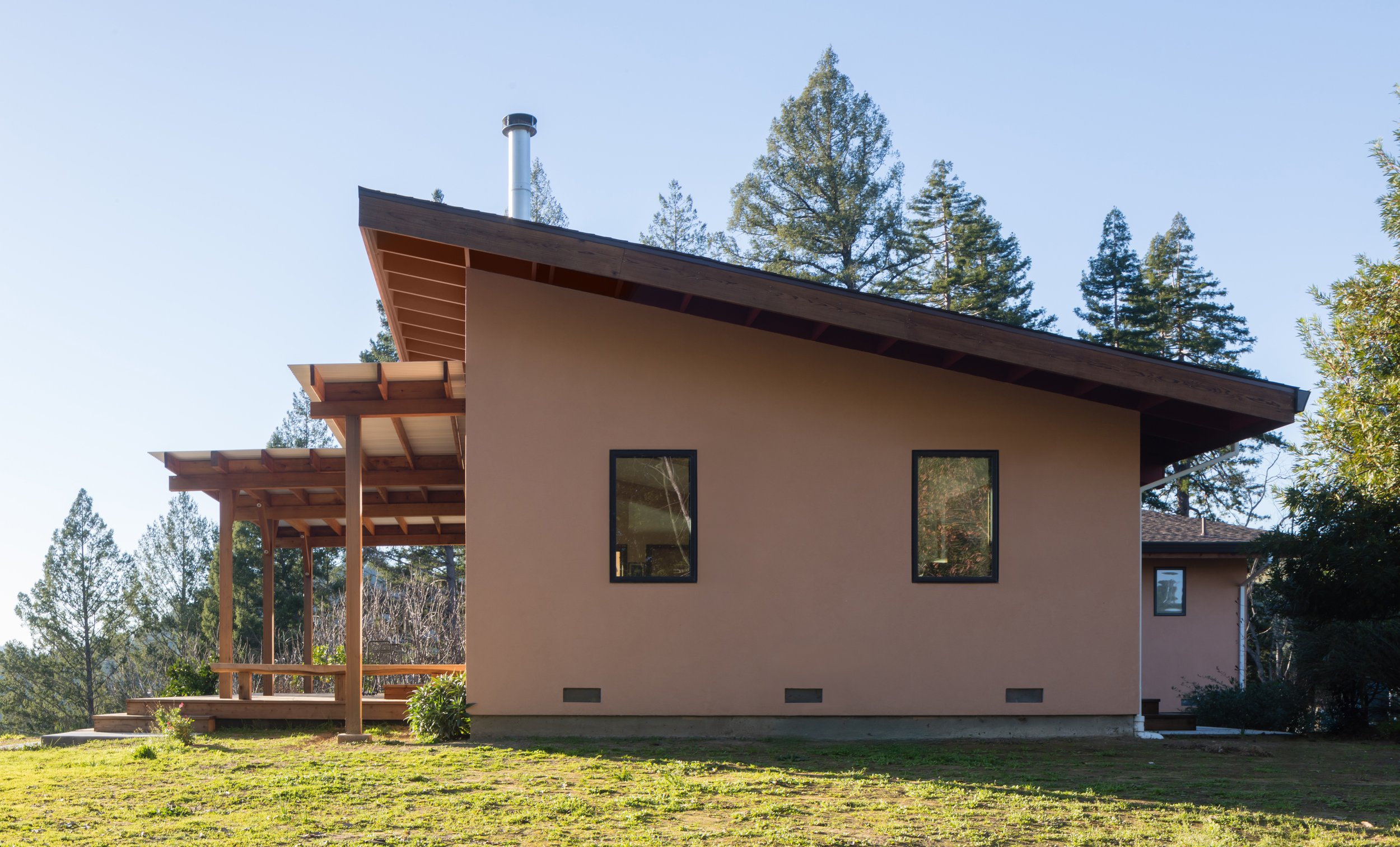 Side view of a modern house with beige walls, large windows, a wooden deck, and a sloped roof, surrounded by trees and grass under a clear blue sky.