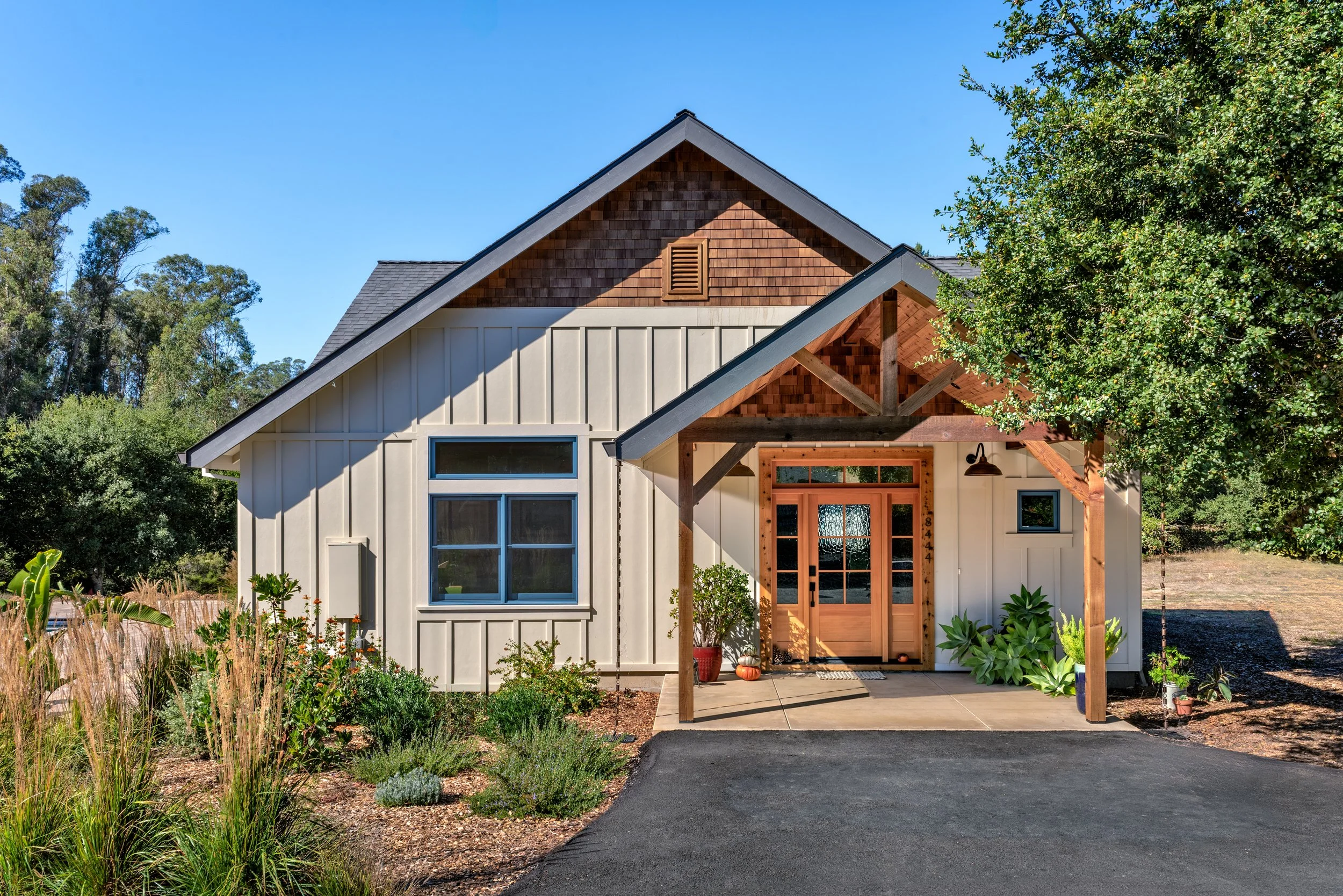 Front of a modern house with white siding, wooden accents, a gabled roof, and a small porch with potted plants, surrounded by landscaping and trees under a clear blue sky.