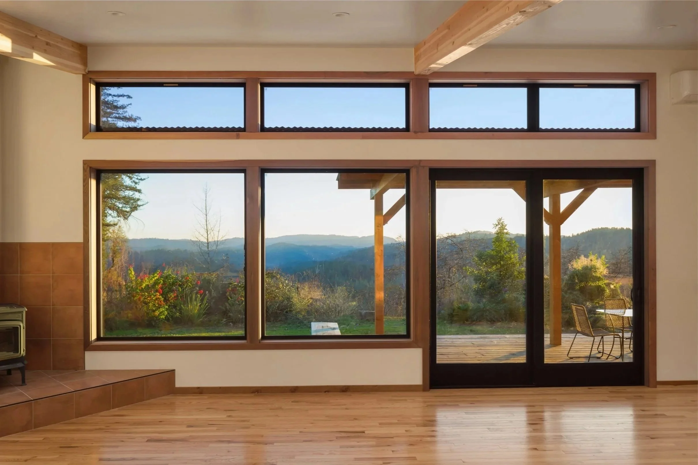Interior view of a room with large windows showing a scenic landscape of trees, mountains, and a blue sky, with a wooden deck outside and wooden beams inside.