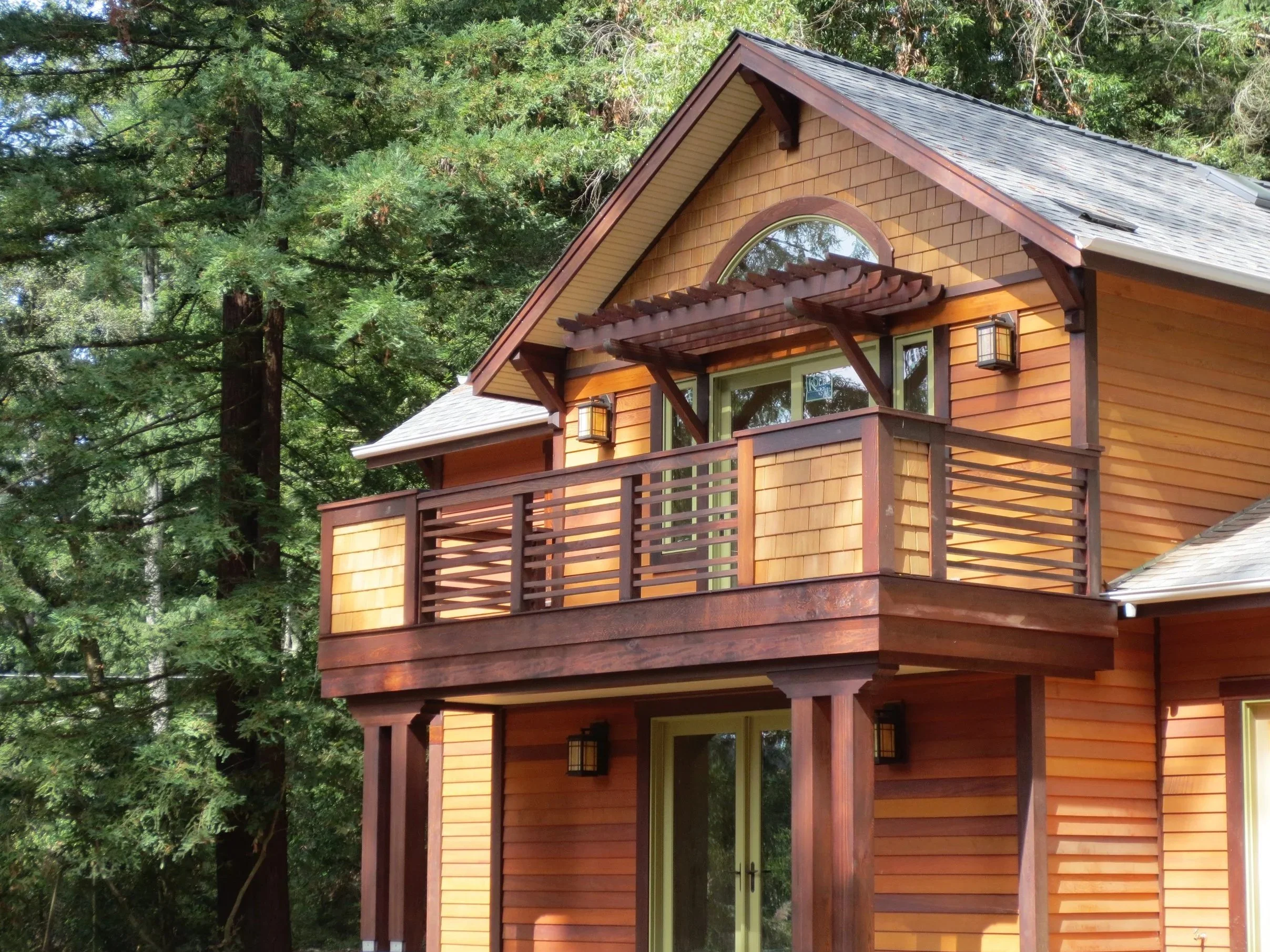 A two-story house in Marin with wood siding, a balcony, and a steep gabled roof surrounded by tall green trees.
