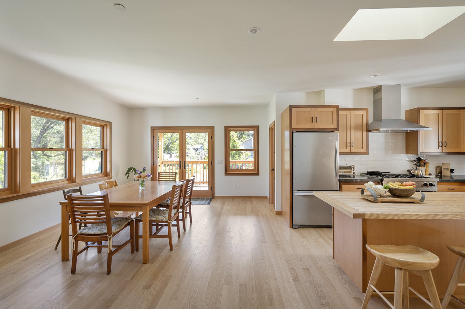 Bright open kitchen and dining area with wooden furniture, large windows, and a sliding glass door leading to a small porch, with light-colored hardwood floors and natural wood cabinetry.