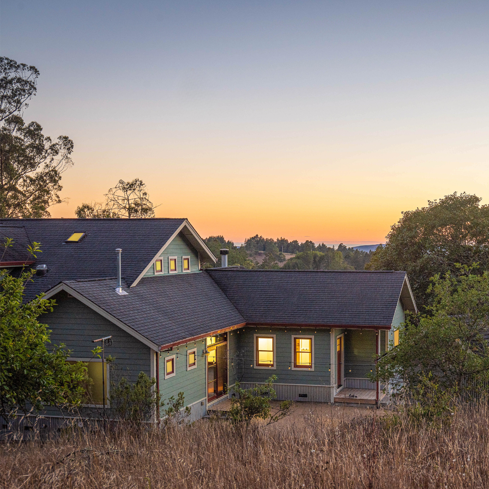 A view of a house with a green exterior and black roof, illuminated from inside, at sunset. The house has multiple large windows and a wrap around deck.