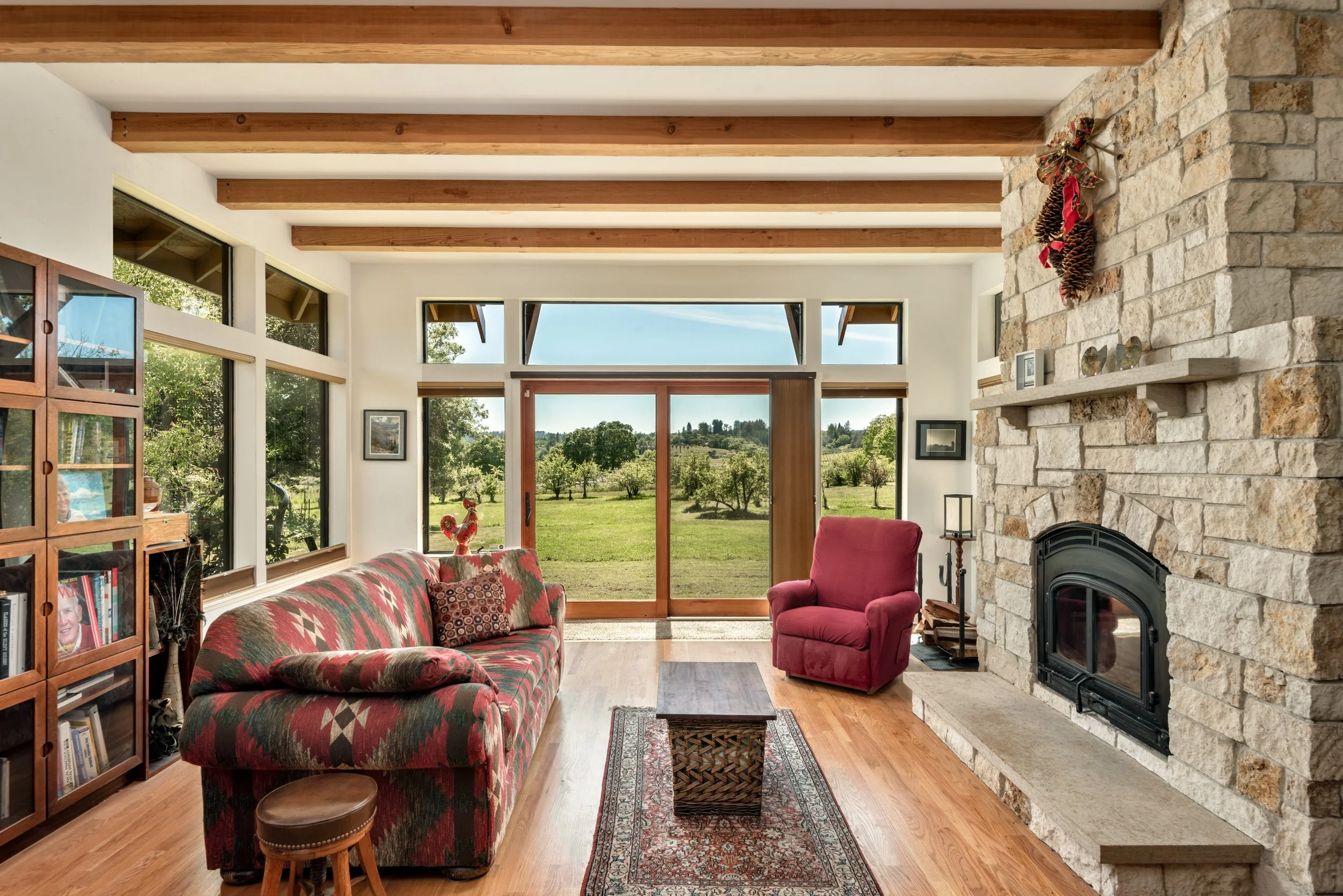 A living room with a stone fireplace, a patterned sofa, a red armchair, large windows showing a green landscape, and decorations including pinecones and holiday ribbons.