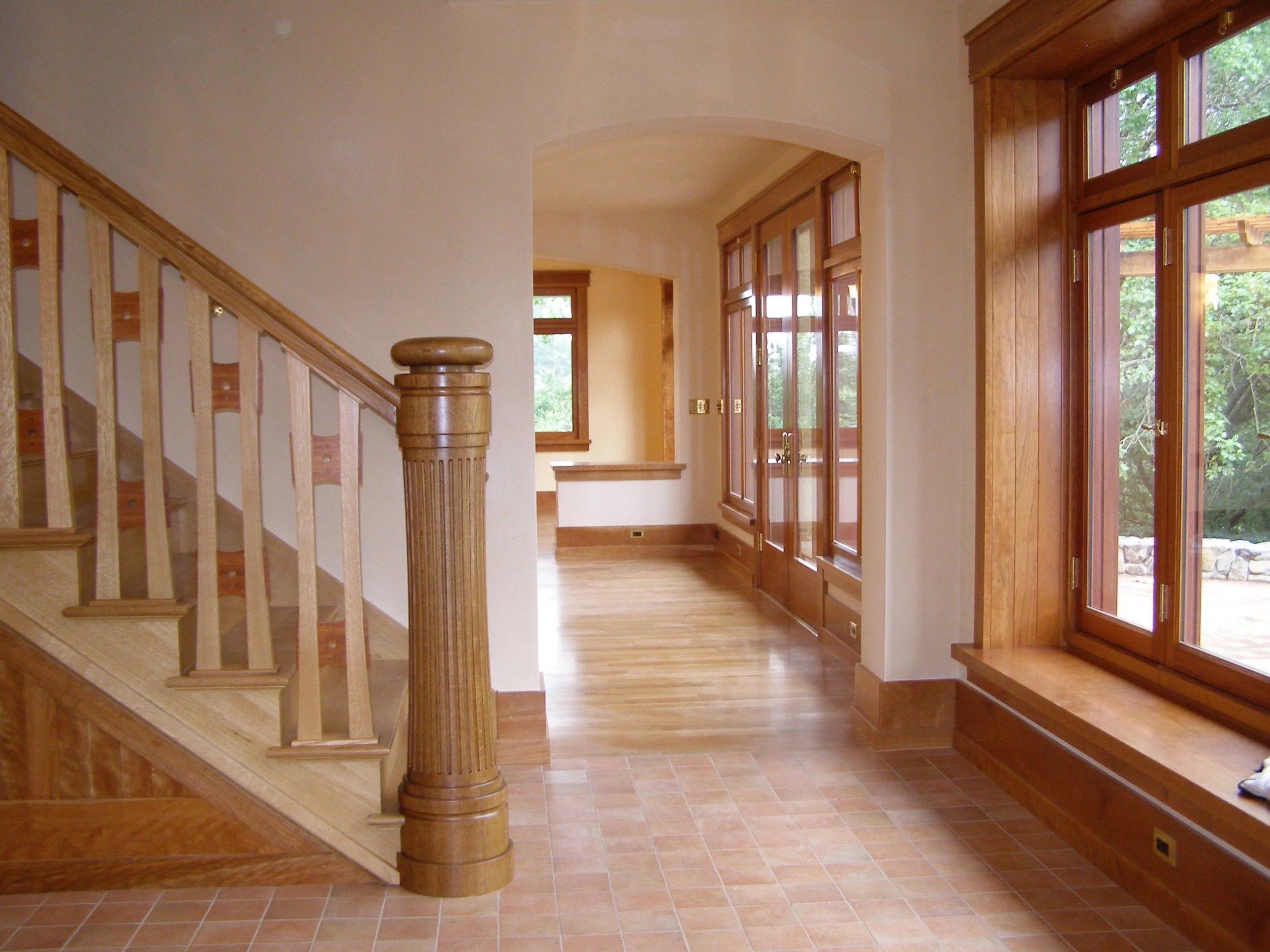 Interior of a house with wooden accents, large windows, a staircase with a wooden railing, and a tiled floor.