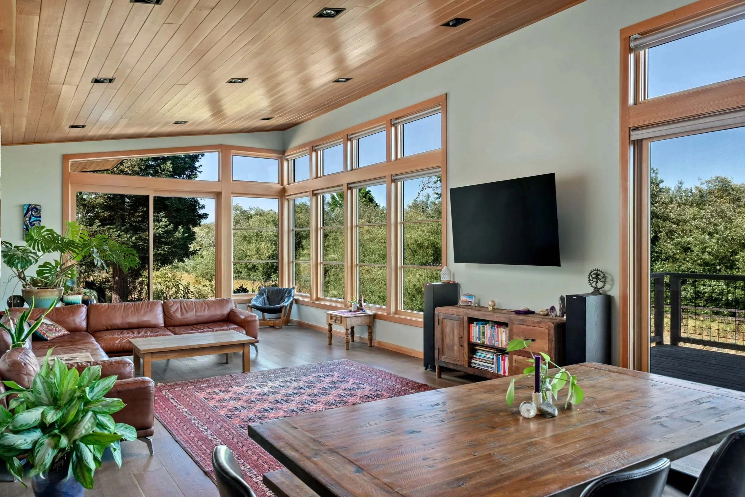 Living room with large windows showing trees outside, leather sofa, wooden coffee table, area rug, potted plants, wooden media console with books, flat-screen TV on the wall, and a wooden dining table.