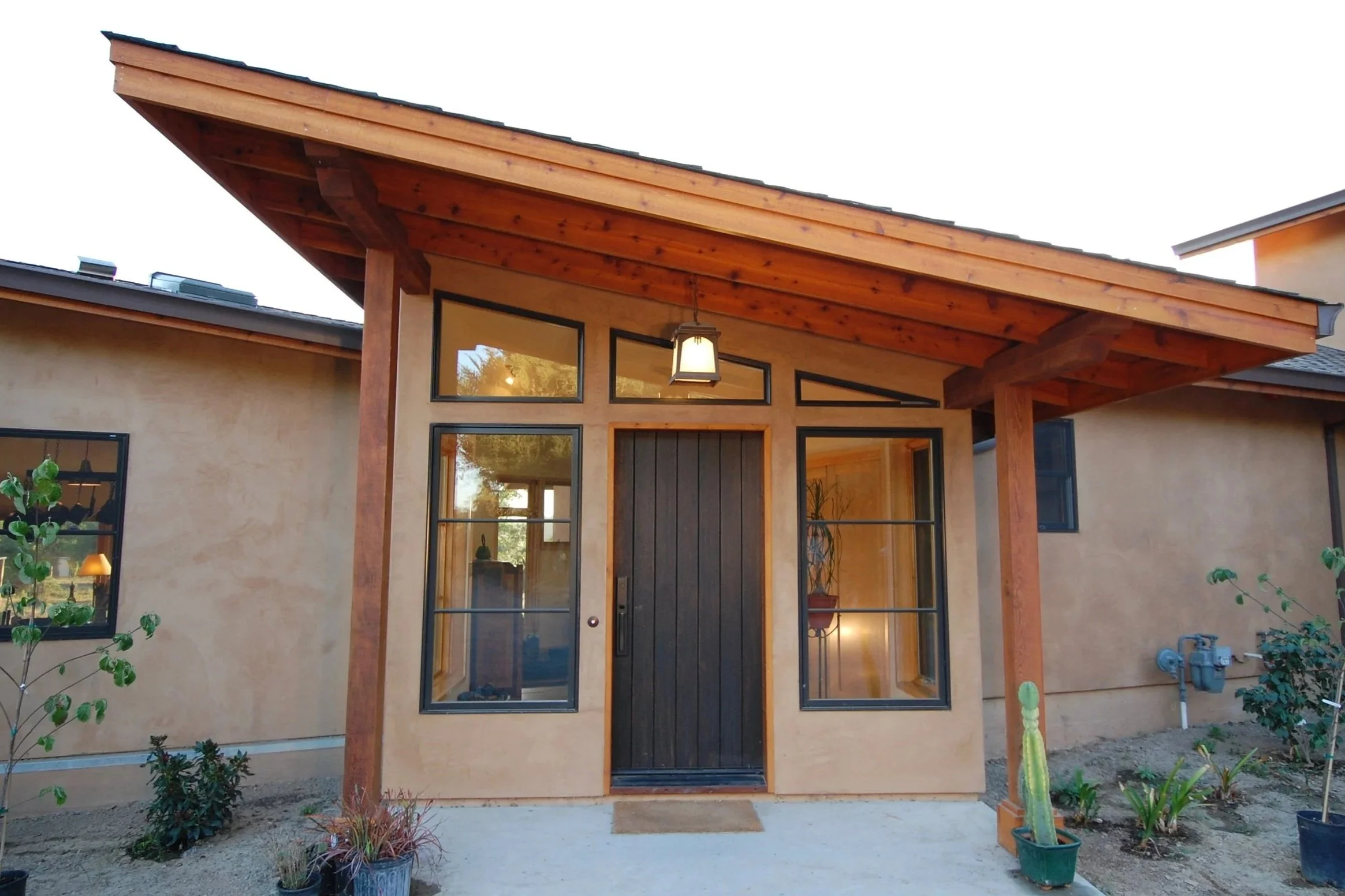 Front view of a modern house with a wooden overhang, large glass windows, and a black front door, surrounded by potted plants and garden plants.
