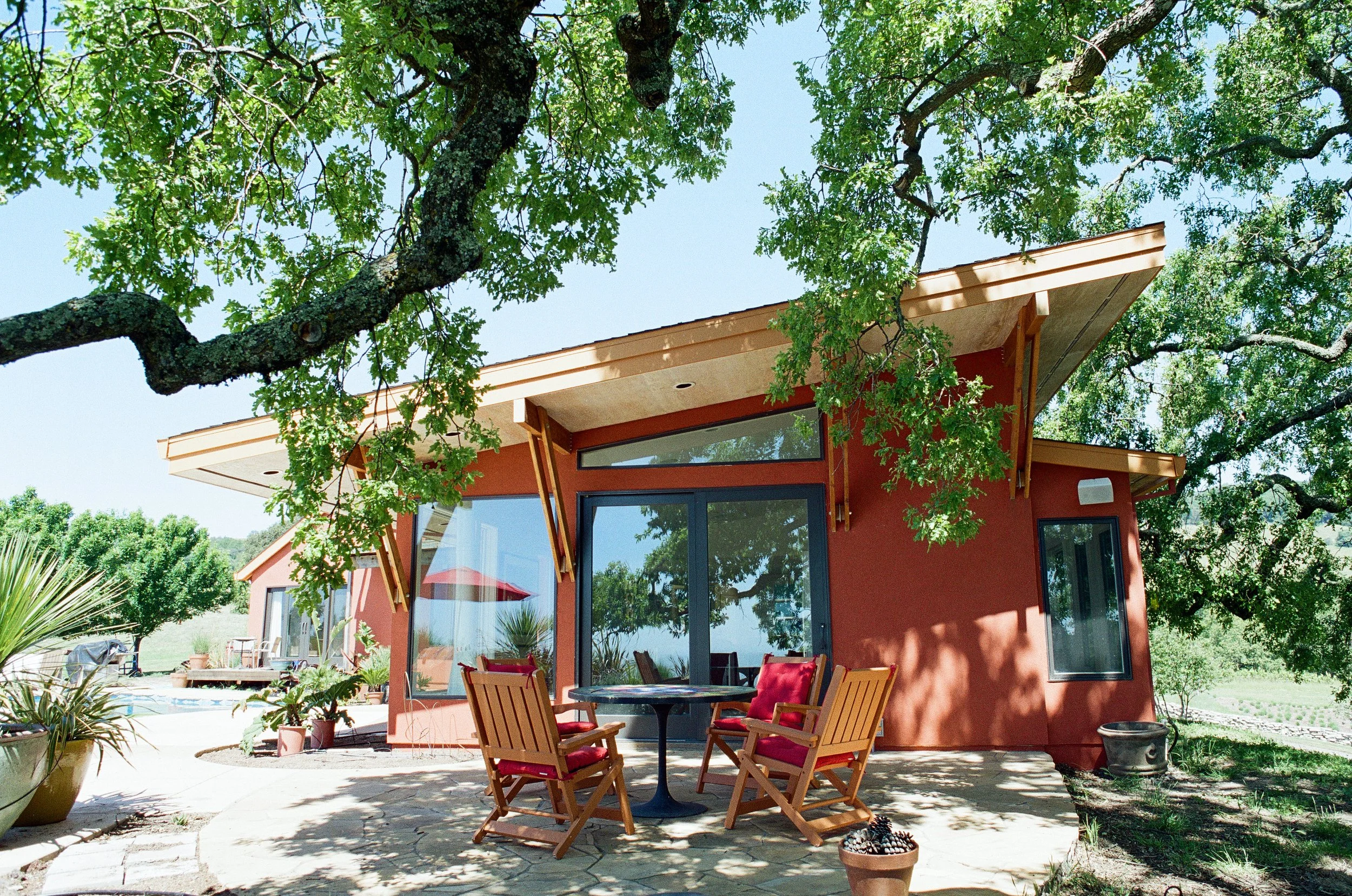 A backyard patio with a round table and four wooden chairs with red cushions, surrounded by potted plants, trees, and a modern house with large windows and an overhanging roof, under a clear blue sky.