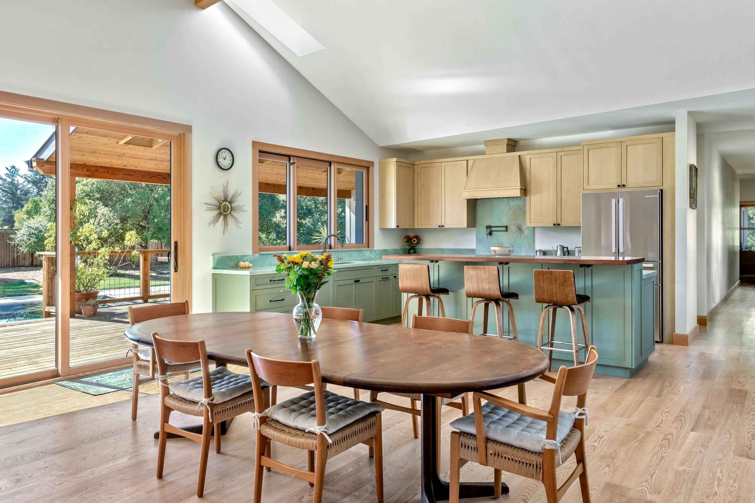 Bright kitchen and dining area with wooden flooring, a round dining table with chairs, a kitchen with light wood cabinets, a green island, and sliding glass doors opening to an outdoor deck with greenery.