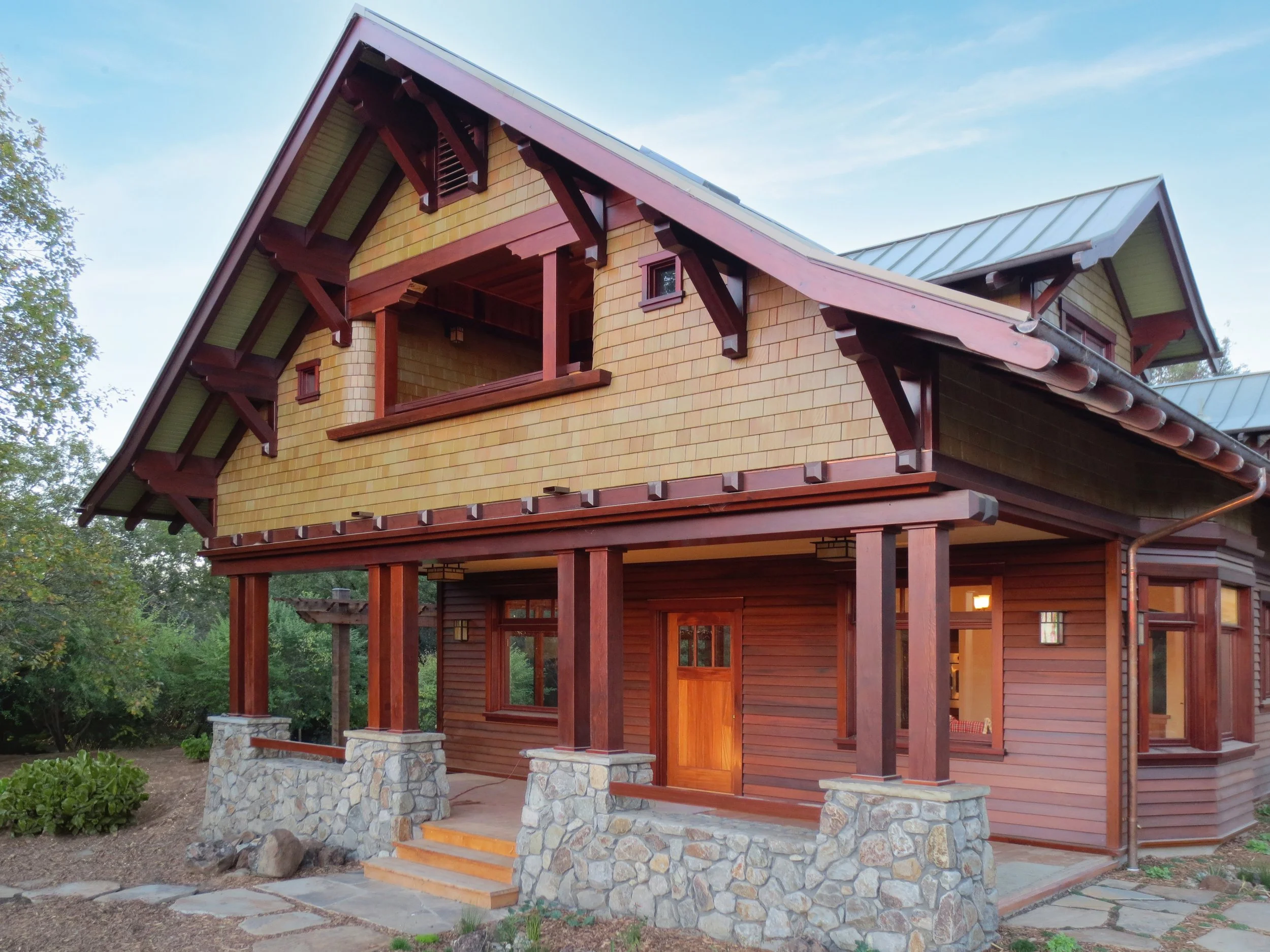 A two-story house with a sloped metal roof, wooden siding, and stone foundation supports. There is a small front porch with stone pillars and steps leading up to a wooden door. The windows are framed in wood, and the house is surrounded by a garden and trees.