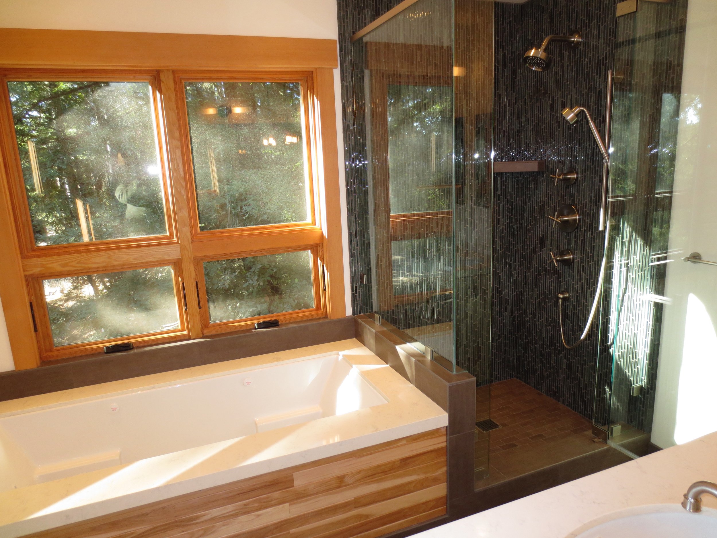 Interior view of a bathroom featuring a large tub with a beige marble surround, a black tiled shower with glass enclosure, multiple showerheads, and a wooden picture window with a view of greenery outside.