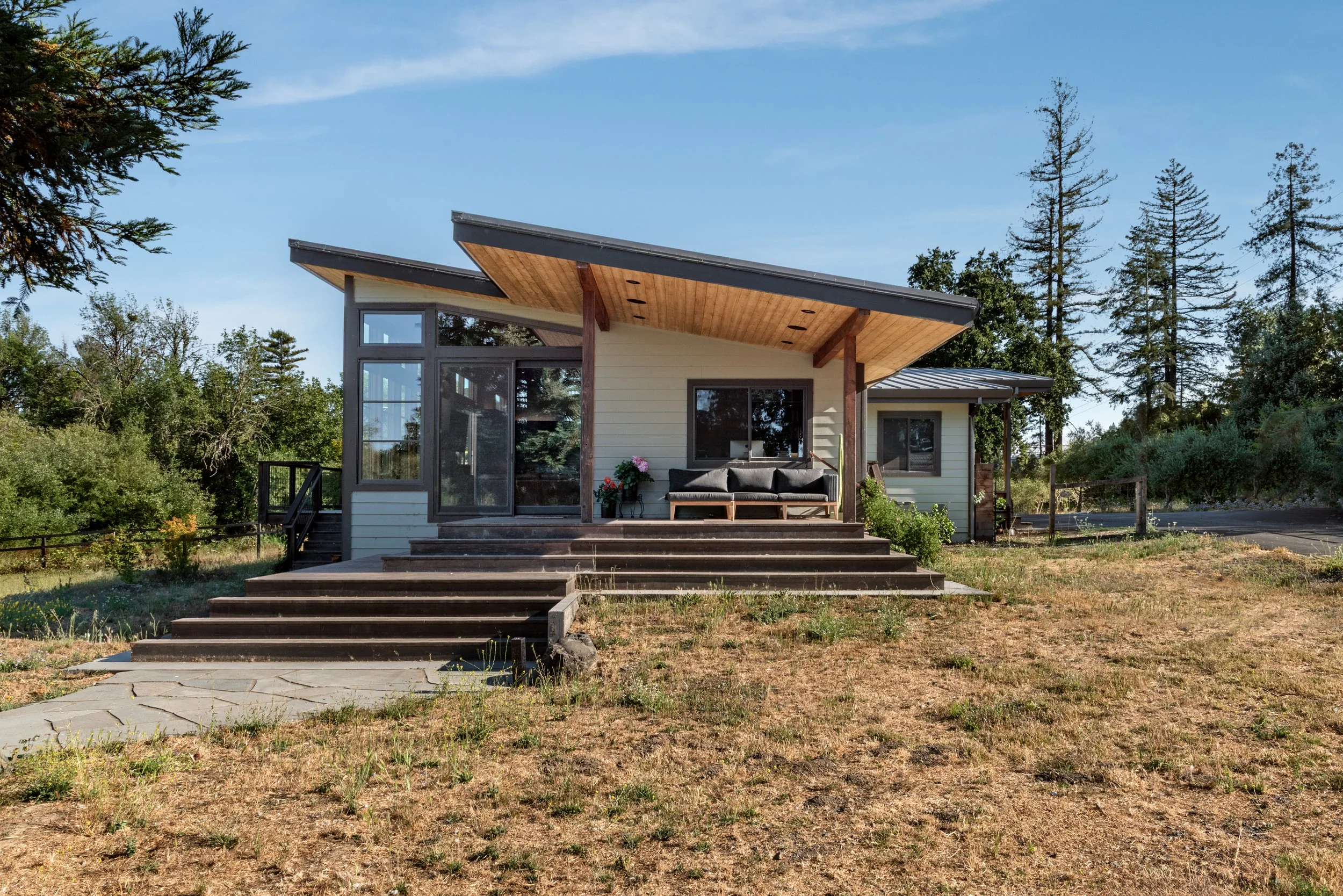Modern house with large windows, sloped roof, and outdoor seating area, set in a wooded area under a clear blue sky.