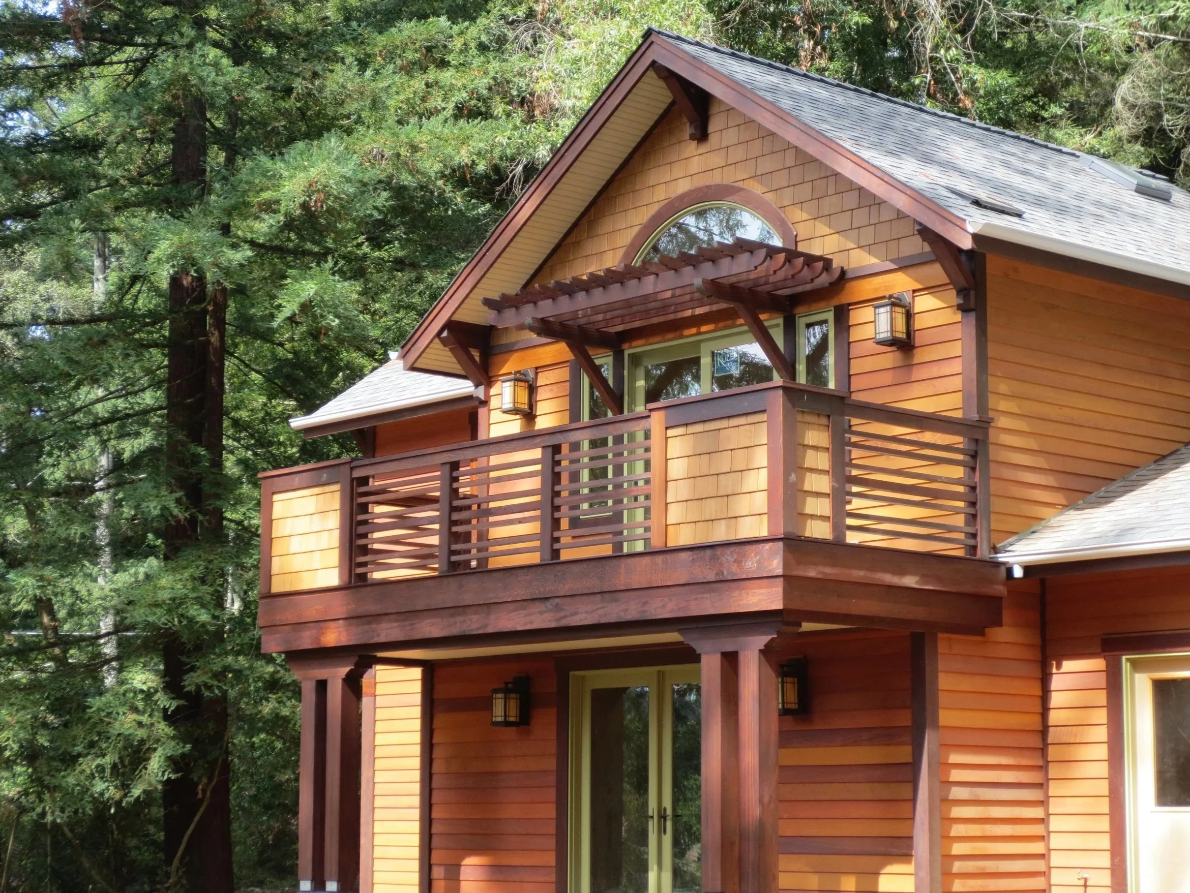 A two-story wooden house with shingle siding, a balcony with wooden railing, and an arched window, surrounded by tall pine trees.