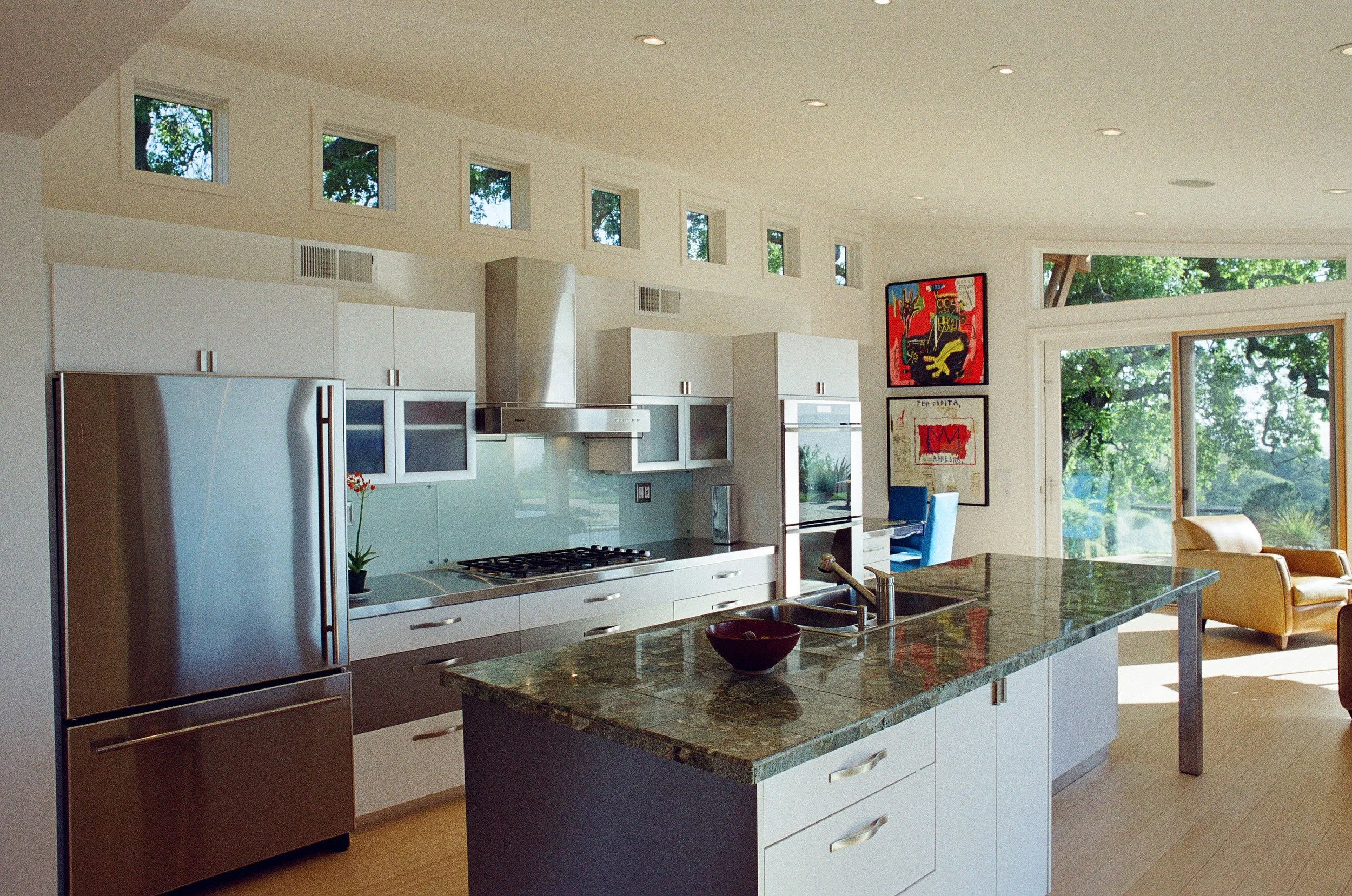 Modern kitchen with white cabinetry, stainless steel appliances, granite island, and large windows letting in natural light, with artwork on the wall and outdoor greenery visible.