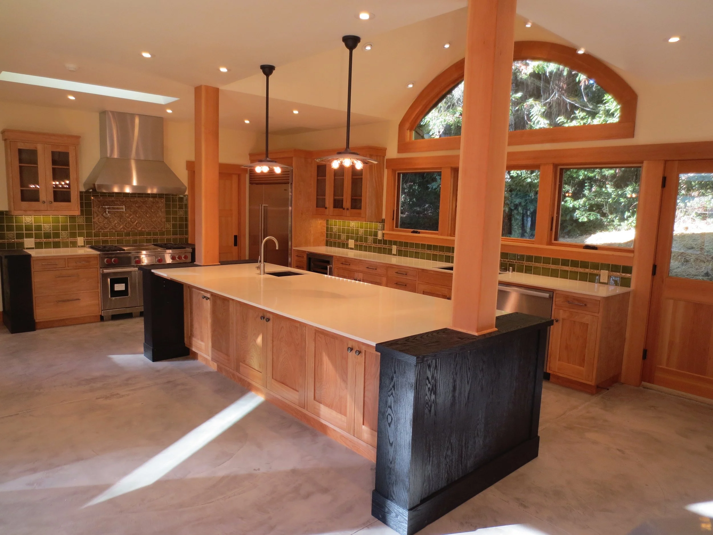 A spacious kitchen with wooden cabinets, green tiled backsplash, and large windows showing trees outside. It features a central island with a sink and modern appliances.