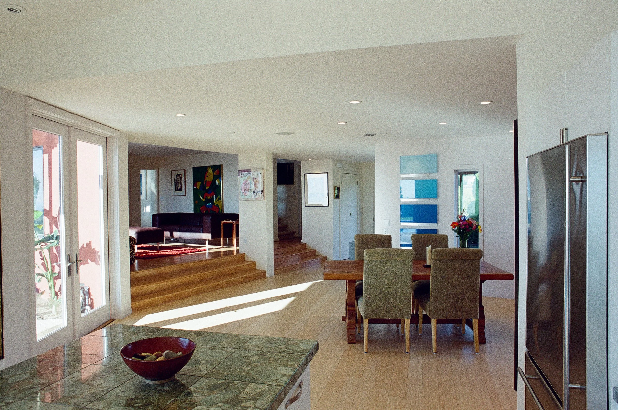 Interior view of a modern home with sliding glass doors, a kitchen island with a red bowl, a dining table with upholstered chairs, and artwork on the walls.