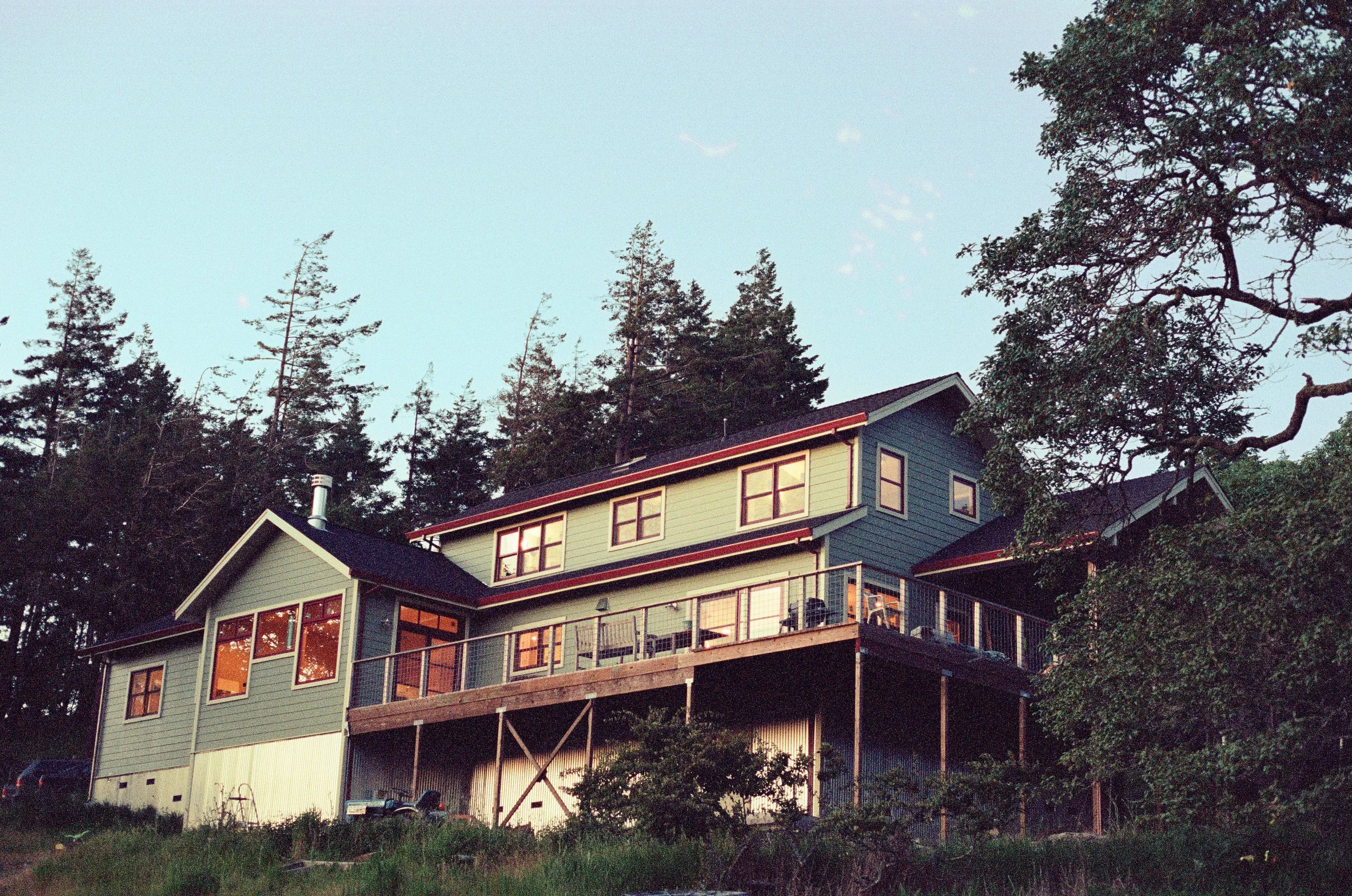 Two-story house with large windows and balcony, situated on a hillside surrounded by trees, with the sky above.