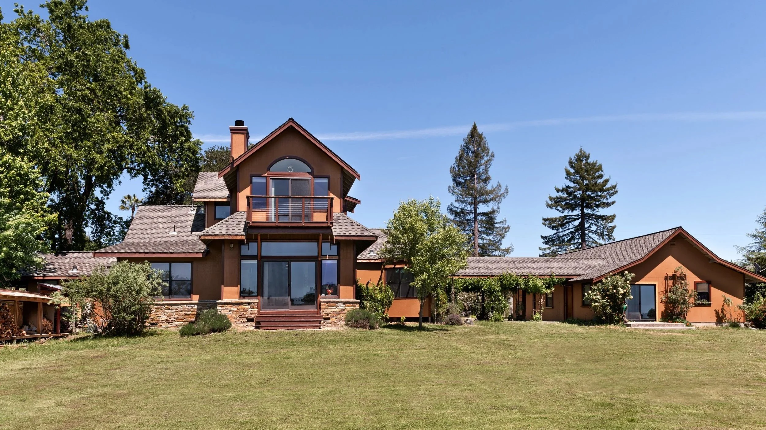 A large two-story house with a brown exterior, multiple windows, a balcony, and a sloped roof surrounded by green trees and a lawn under a clear blue sky.