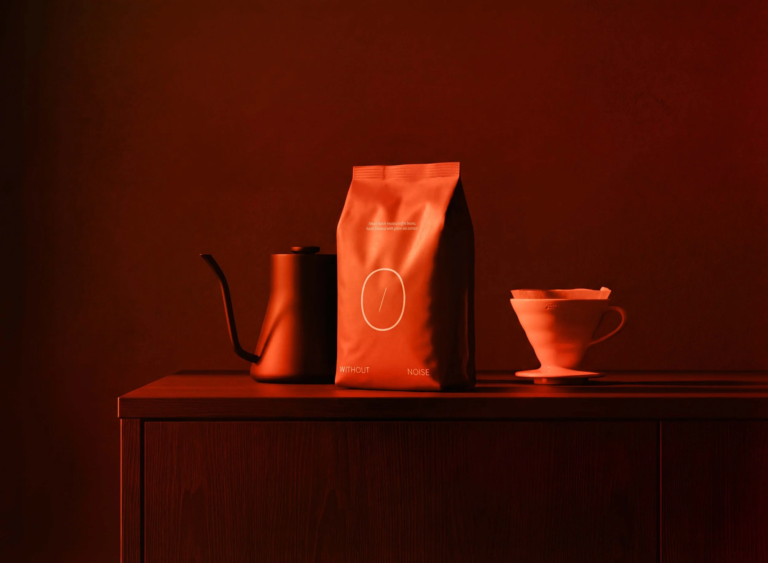A black coffee pot, a bag of coffee, and a cup on a wooden surface against a dark background with red lighting.