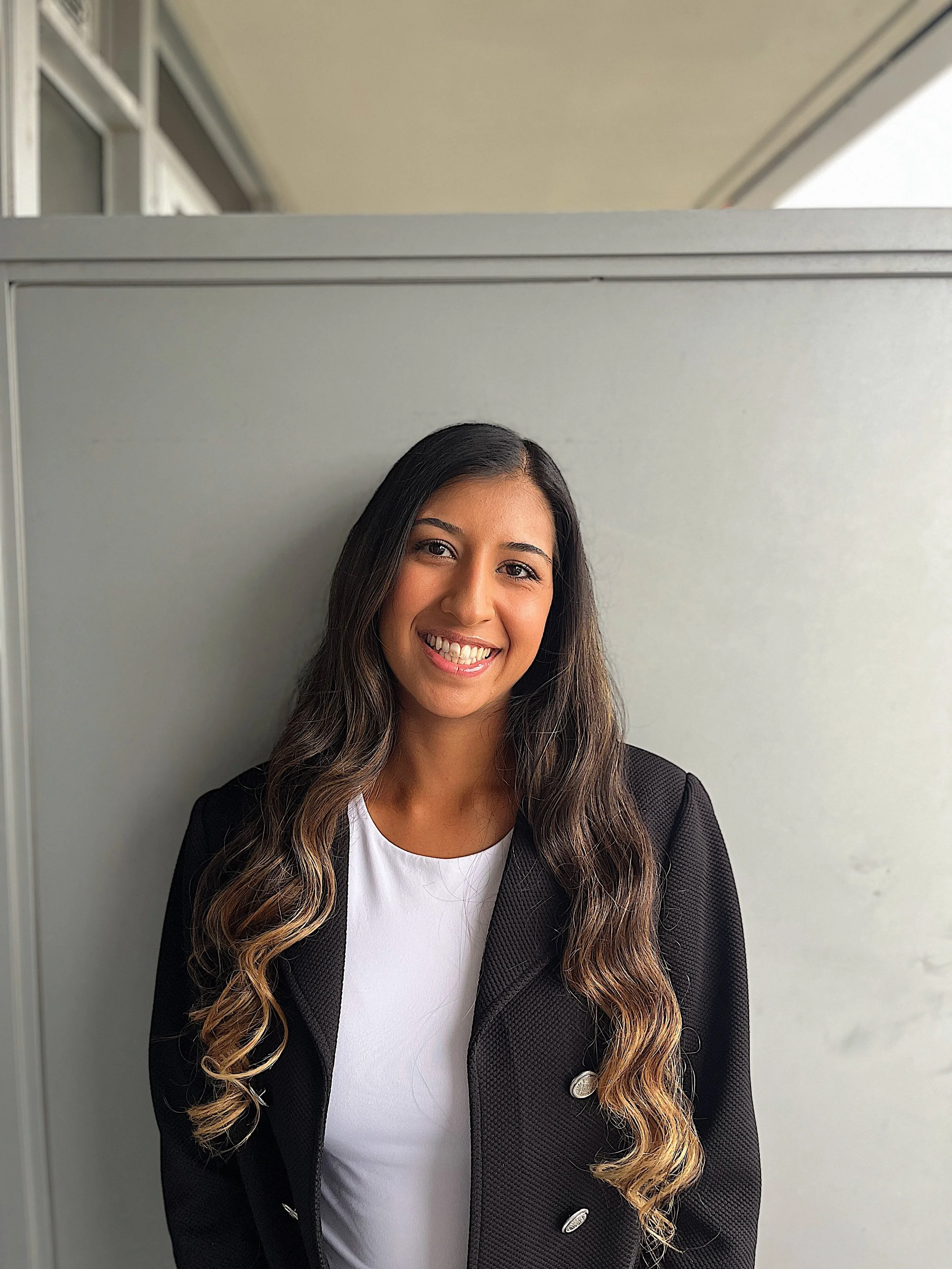 A young woman with long wavy hair smiling, wearing a black blazer over a white top, standing against a gray wall near windows.