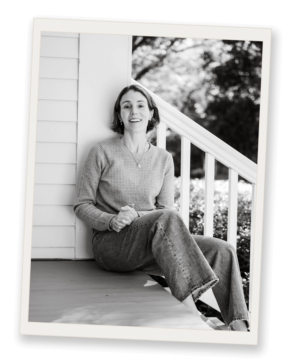 Black and white photo of smiling woman sitting on a porch step, leaning against a wall, with a railing and trees in the background.