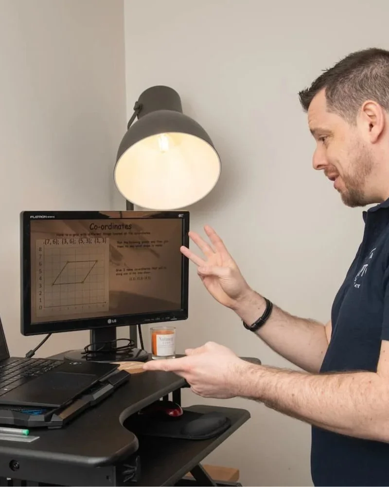 A man in a dark polo shirt demonstrating a 3D coordinate graph on a computer monitor in a home office, with a desk lamp above and a notebook on the desk.