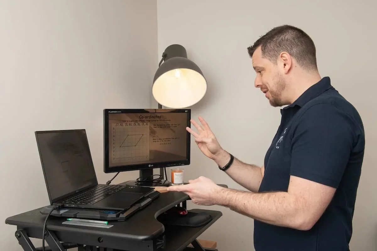 Nick Buckley, Private Tutor in Driffield and East Yorkshire at PTO Private Tuition Online standing at a desk with a laptop and a monitor, gesturing with his right hand while looking at the monitor displaying a graph and text about coordinates.