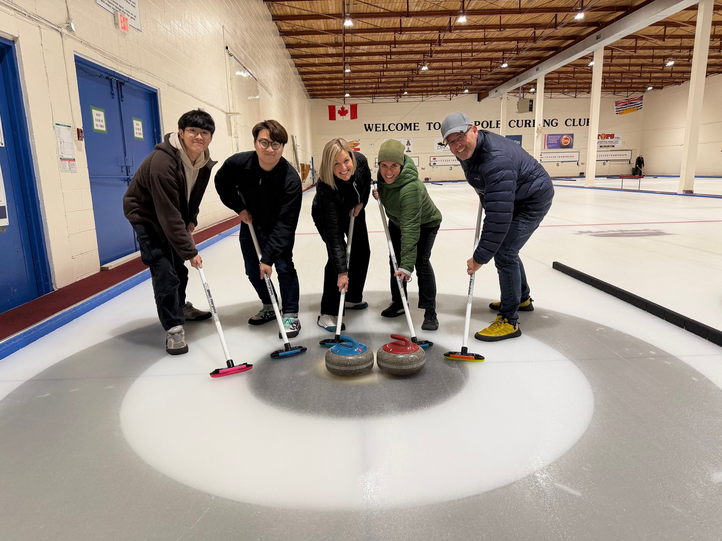 Five people are curling on an indoor ice rink, all holding curling brooms and positioned around curling stones. They are smiling and bent forward in a curling stance. The background shows a wall with a Canadian flag, the words 'Welcome to Curling Clu