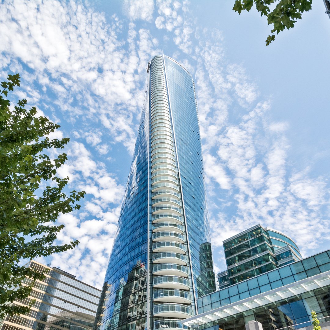 A modern glass skyscraper reaching into a partly cloudy blue sky, with surrounding buildings and green trees at the base.