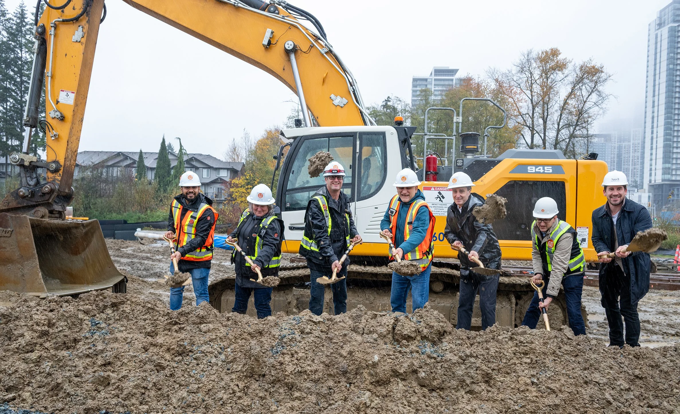 Group of construction workers wearing hard hats and safety vests standing in front of a large excavator, holding shovels, at a construction site with buildings and trees in the background.