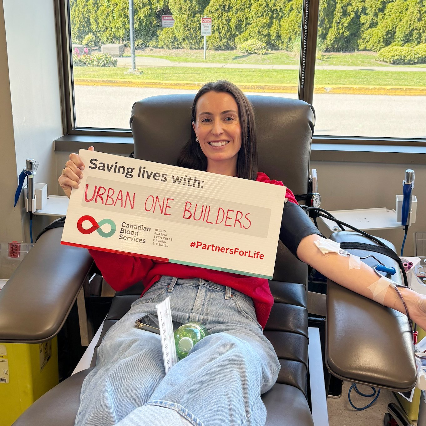 A woman sitting in a blood donation chair, smiling, holding a sign that reads 'Saving lives with: URBAN ONE BUILDERS' and '#PartnersForLife'. She has a needle in her arm connected to a blood collection tube. The background shows a window with a view 