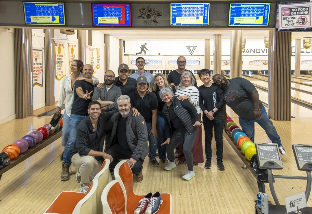 A group of fifteen people posing together at a bowling alley, with bowling balls on racks and digital score screens overhead.