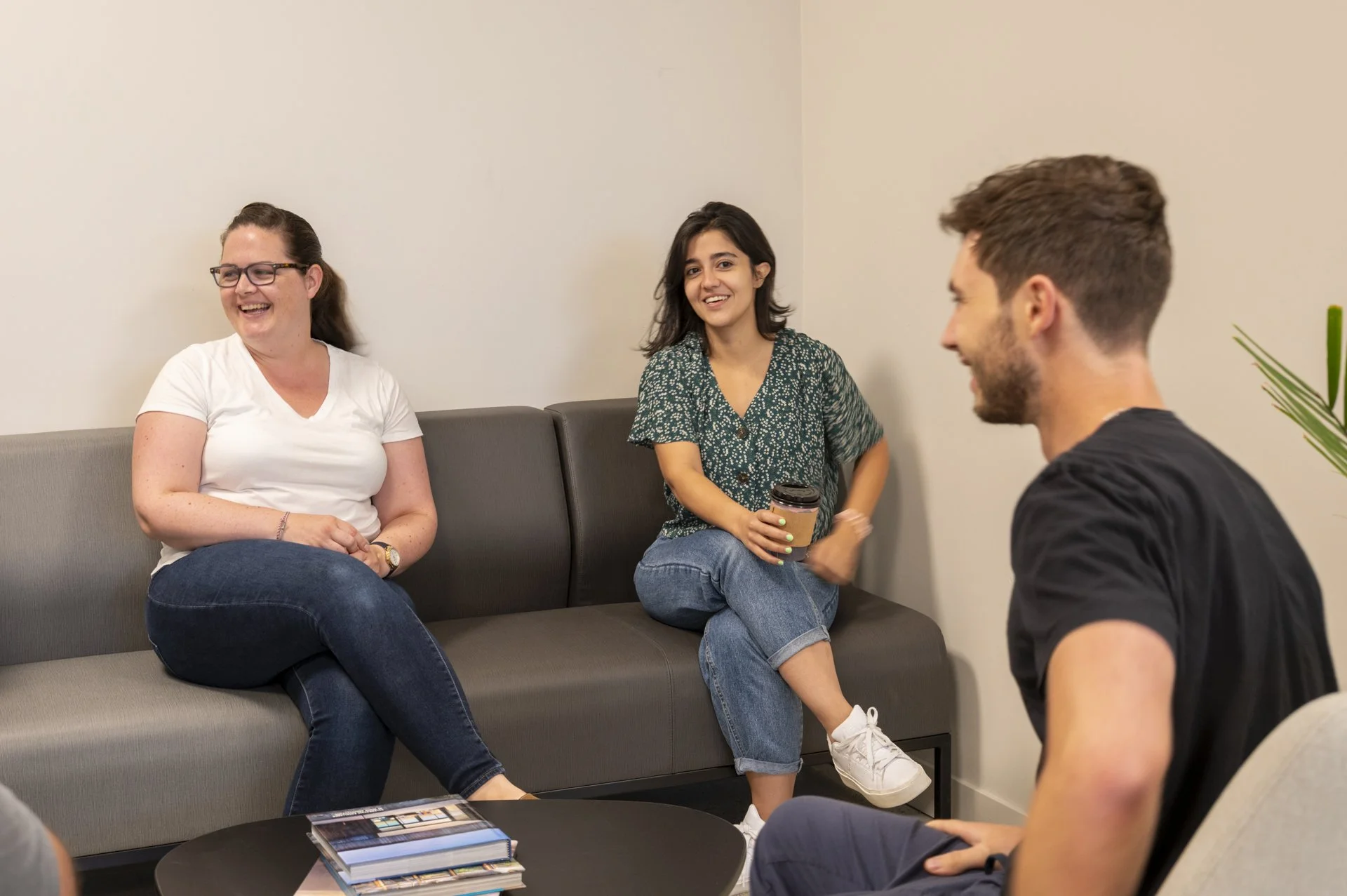 Three young adults, two women and a man, sitting and chatting in a casual setting, with a table holding magazines in front of them. The women are smiling and the man is talking, with a plain wall and a potted plant in the background.