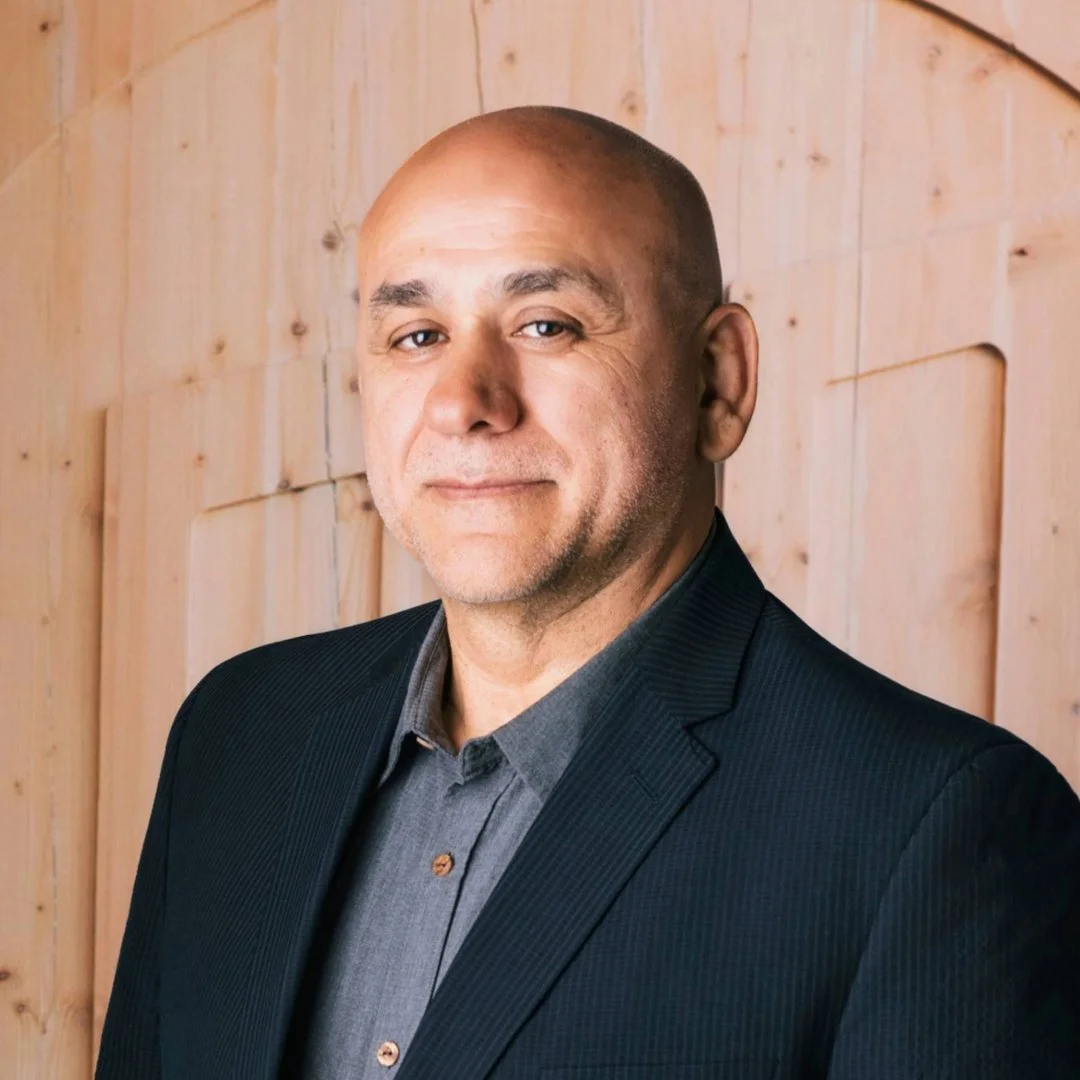 A middle-aged man with a shaved head, wearing a dark suit jacket and gray shirt, stands against a wooden wall background.