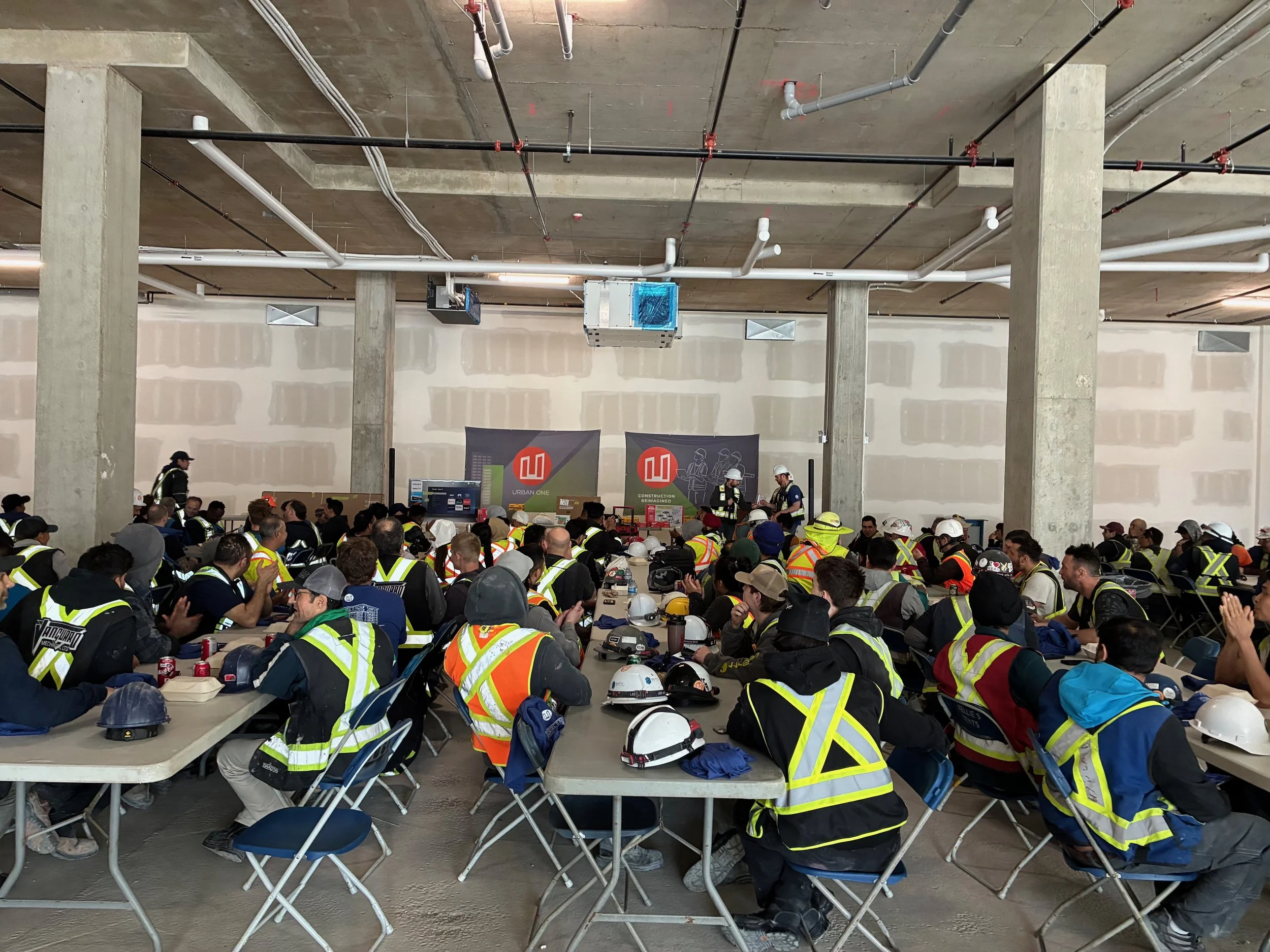 A large group of construction workers wearing safety vests and helmets attending a meeting or presentation inside an unfinished building. The ceiling has exposed pipes and ductwork, and there are banners with construction logos.