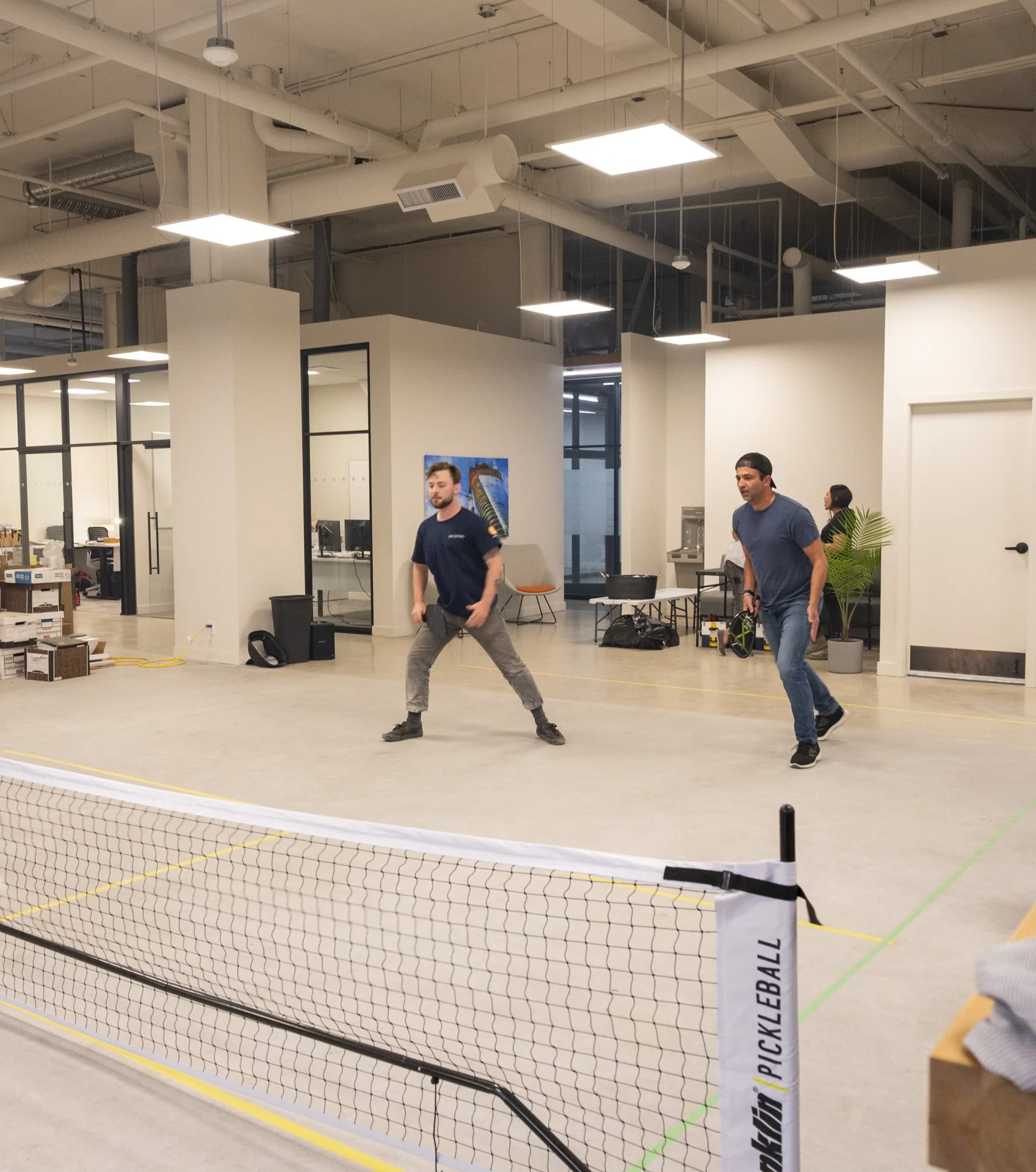 People playing pickleball indoors on a court with a net. The court has markings and barriers around it and some equipment and furniture visible in the background.