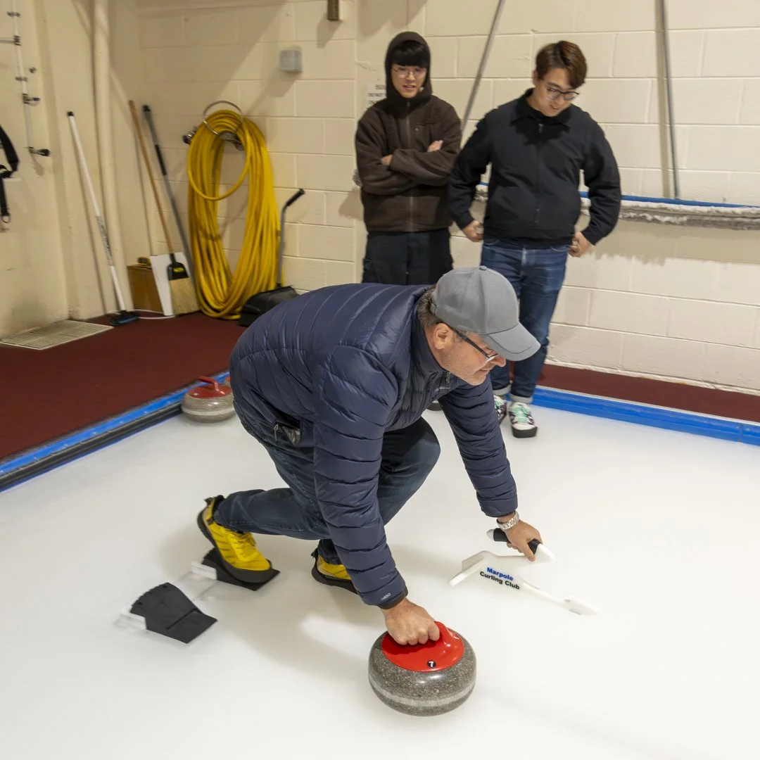 Person preparing to throw a curling stone on an indoor ice rink while two people observe.
