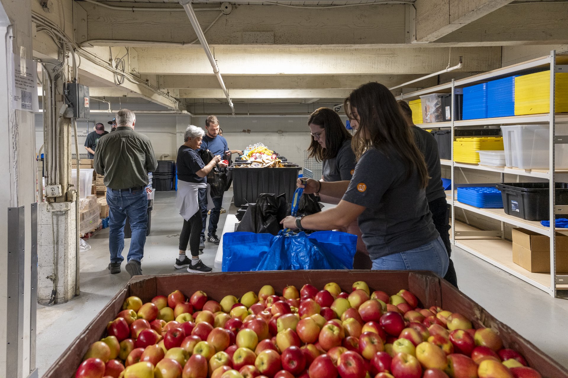 A large crate filled with red and yellow apples in the foreground, with several people in casual clothing and gloves working at a table behind it, sorting and packing apples inside a storage or warehouse space with shelves on the side.