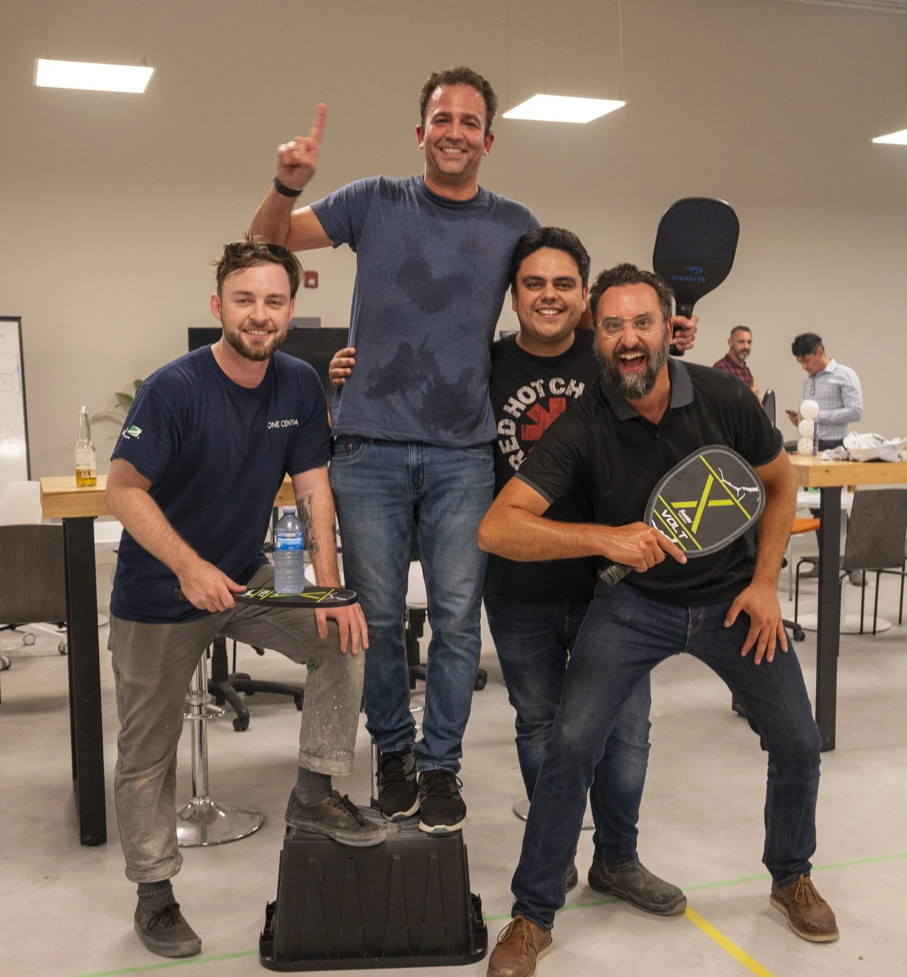 A group of four men smiling in an indoor workspace. One man is standing on a black box, holding a ping pong paddle, while the others are seated or standing appropriately around him. The man on the left has a beard and is wearing a dark blue t-shirt, 