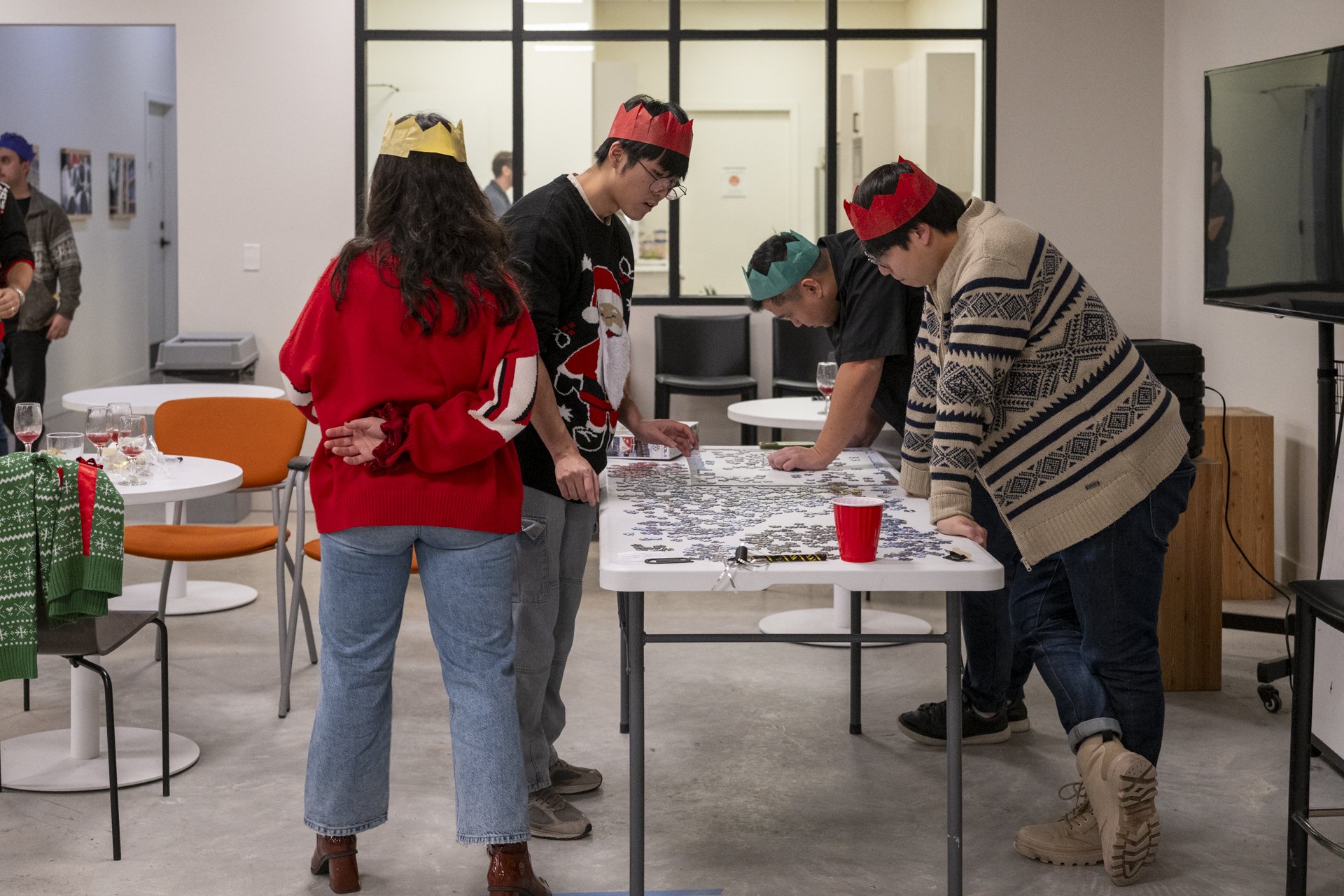Four people wearing Christmas hats working on a jigsaw puzzle at a table in a festive room.