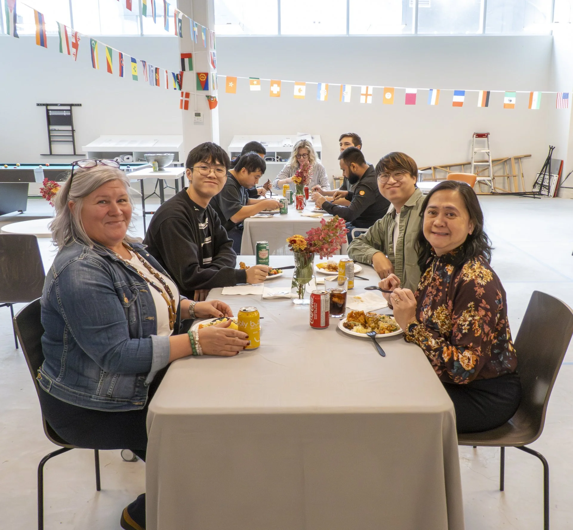 People sitting at a long table covered with a white tablecloth during a meal or gathering, with colorful flags hanging overhead in a bright, spacious room.