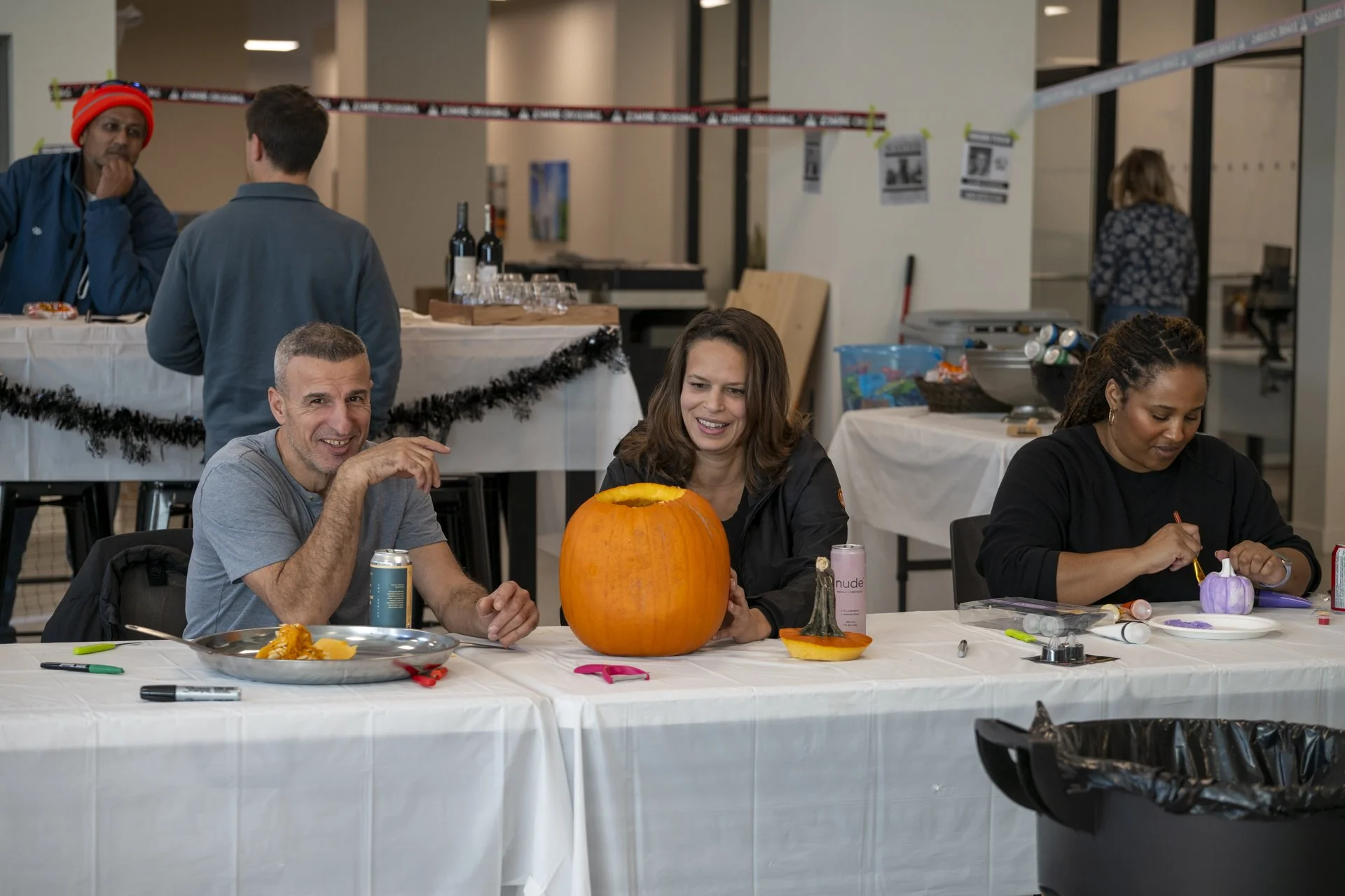 People sitting at a table with a large pumpkin and pumpkin pieces, engaging in a pumpkin decorating activity in an indoor setting.
