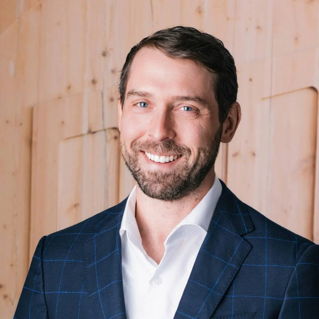 A man with dark hair and a beard, dressed in a white shirt and a dark blue checkered blazer, smiling in front of a wooden wall.