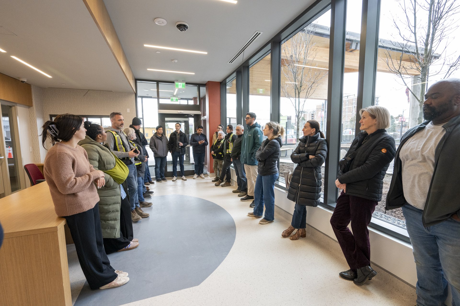 A group of people standing inside a modern building with large floor-to-ceiling windows, listening to a speaker at the front.