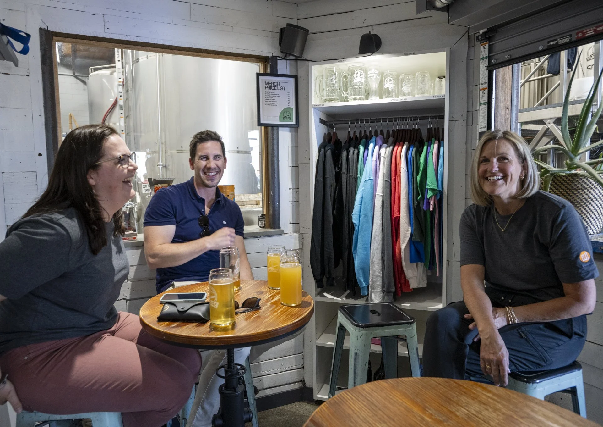 Three women and one man sitting at a round table inside a restaurant or brewery, smiling and laughing, with several glasses of beer on the table, a small cabinet with hanging T-shirts, glass mugs on a shelf, and industrial decor in the background.